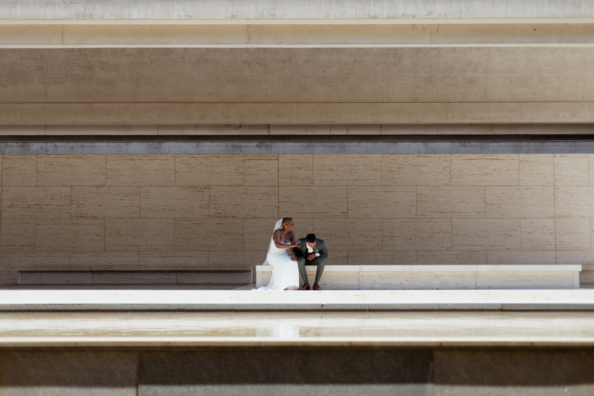 A bride and groom sitting together on a minimalist concrete ledge against a large, beige wall in a modern, architectural setting.
