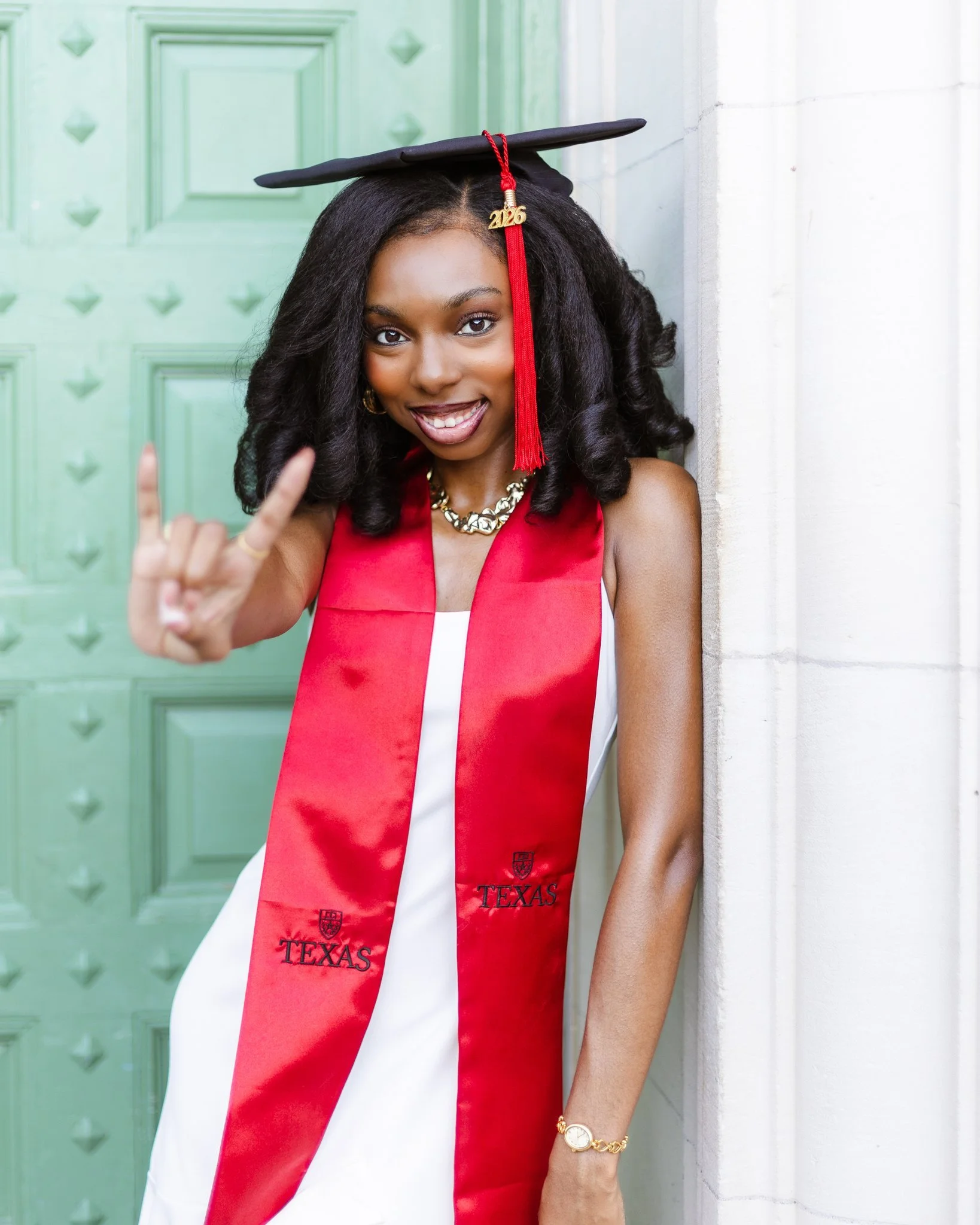 A young woman in her graduation cap and gown, showing an excited expression, making a peace sign with her right hand, standing in front of a green door.