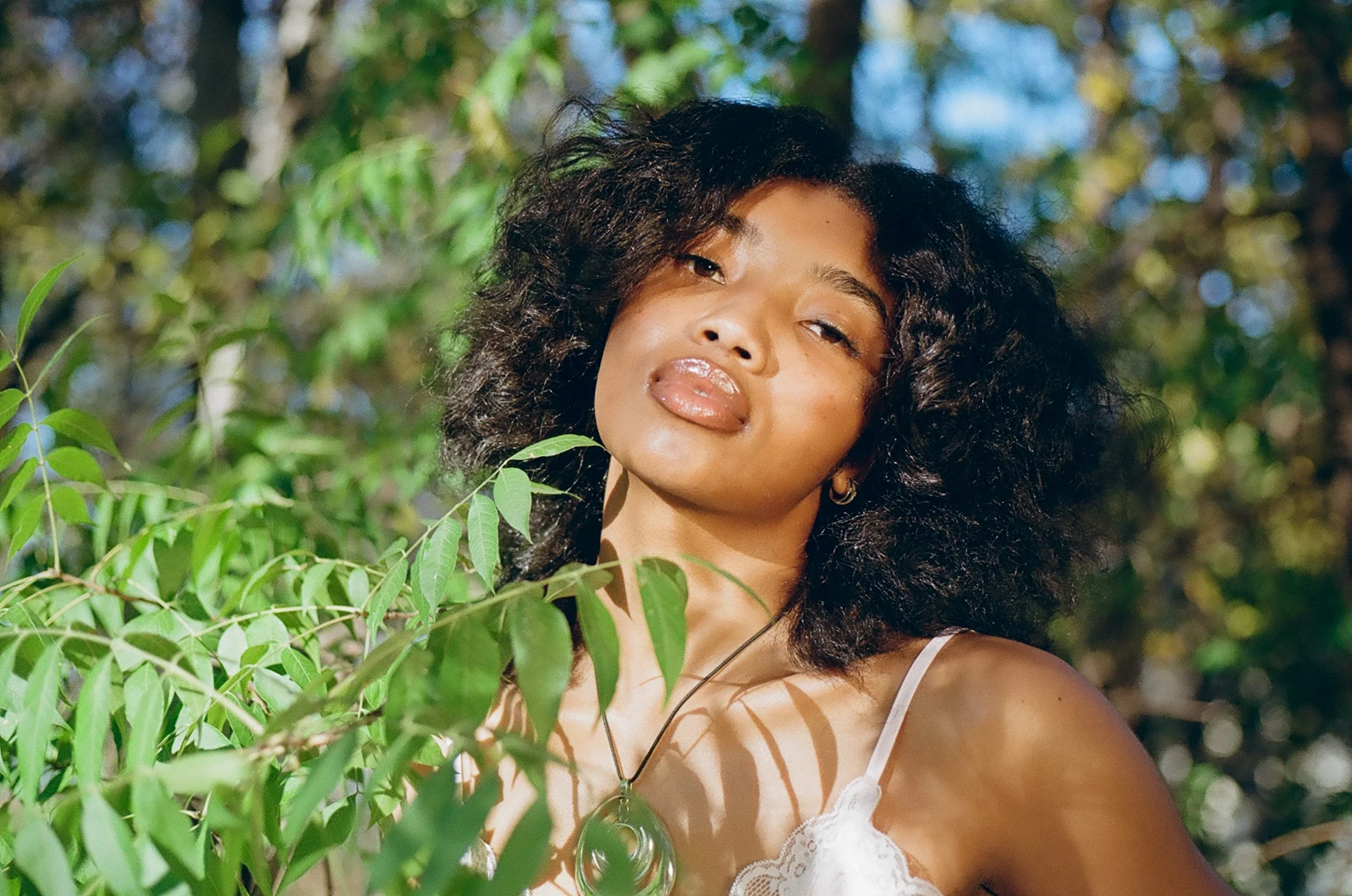 A woman with dark, curly hair stands outdoors among green foliage, wearing a white lace top and a silver necklace with a green stone pendant.