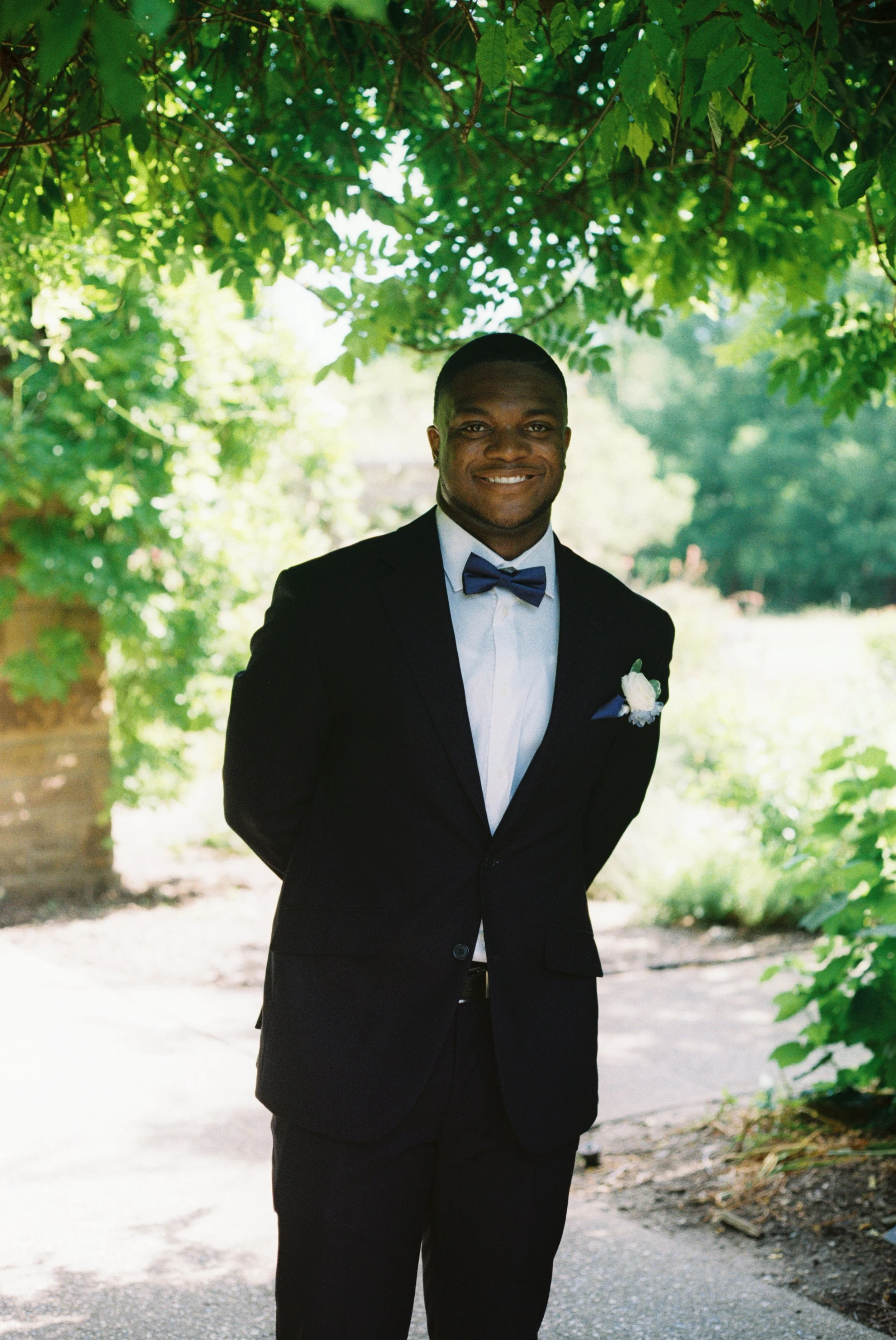 A man wearing a black tuxedo with a bow tie, standing outdoors under green foliage, smiling at the camera.