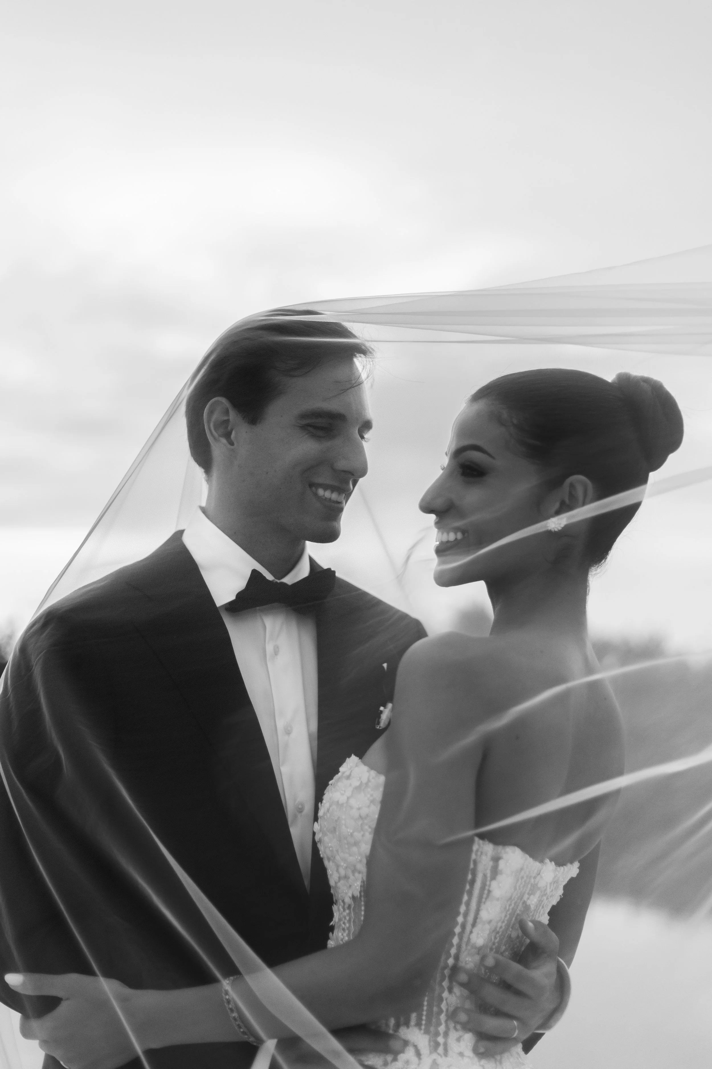 Black-and-white photo of a bride and groom smiling at each other, wrapped in a sheer veil, outdoors.