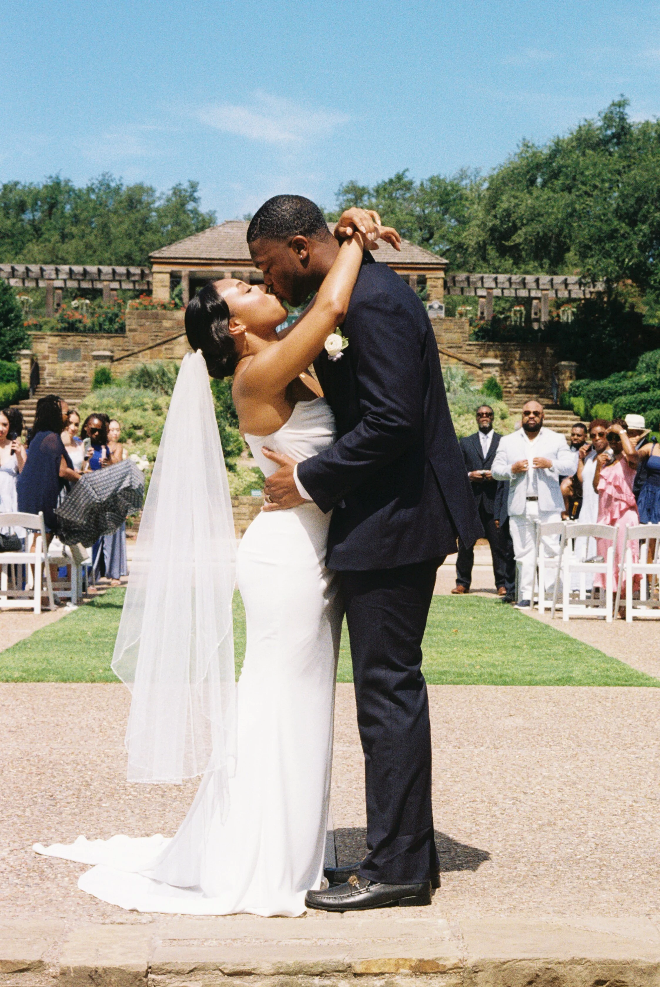 A bride and groom kiss during their outdoor wedding ceremony, with guests watching in the background.