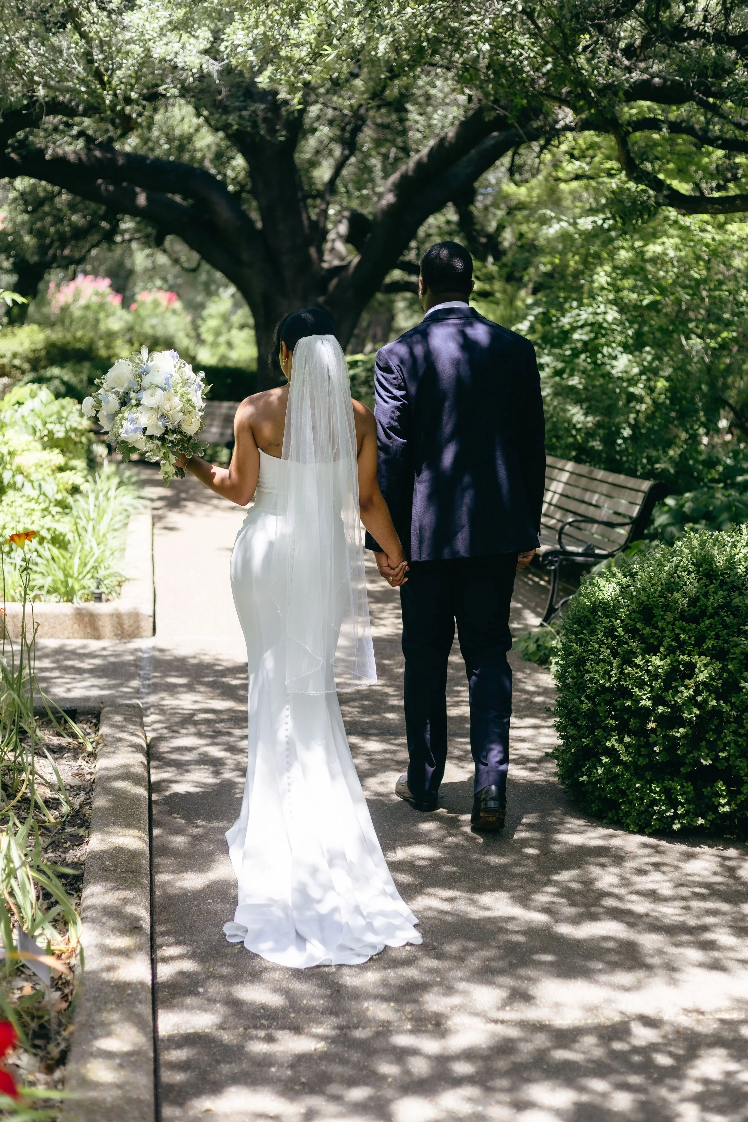 A bride and groom holding hands walking through a lush, green park, with the bride holding a white bouquet, surrounded by trees and foliage.