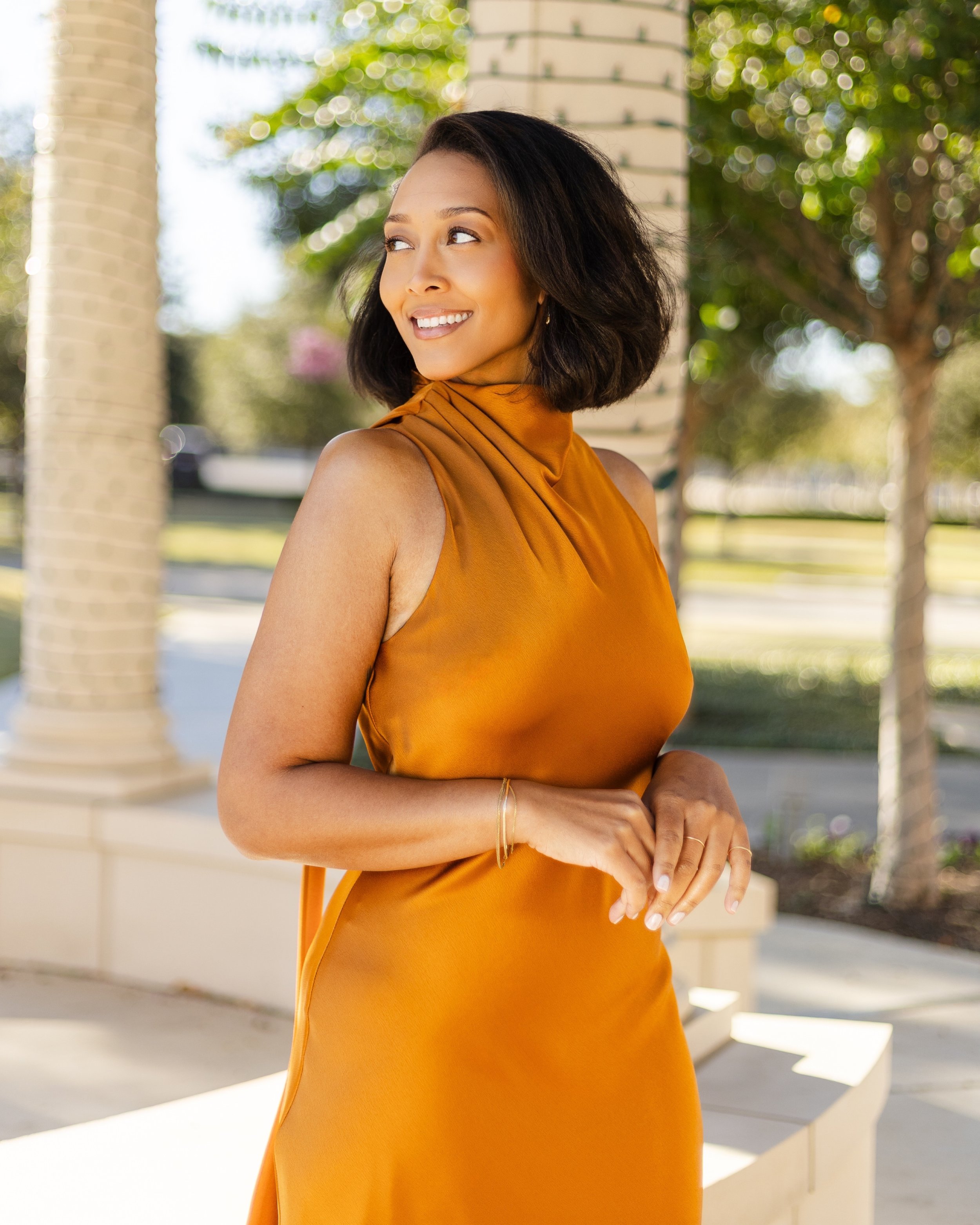 A woman with shoulder-length dark hair, wearing a sleeveless, high-neck, orange satin dress, standing outside near a white column with brick accents, smiling and looking to her left on a sunny day with trees and greenery in the background.