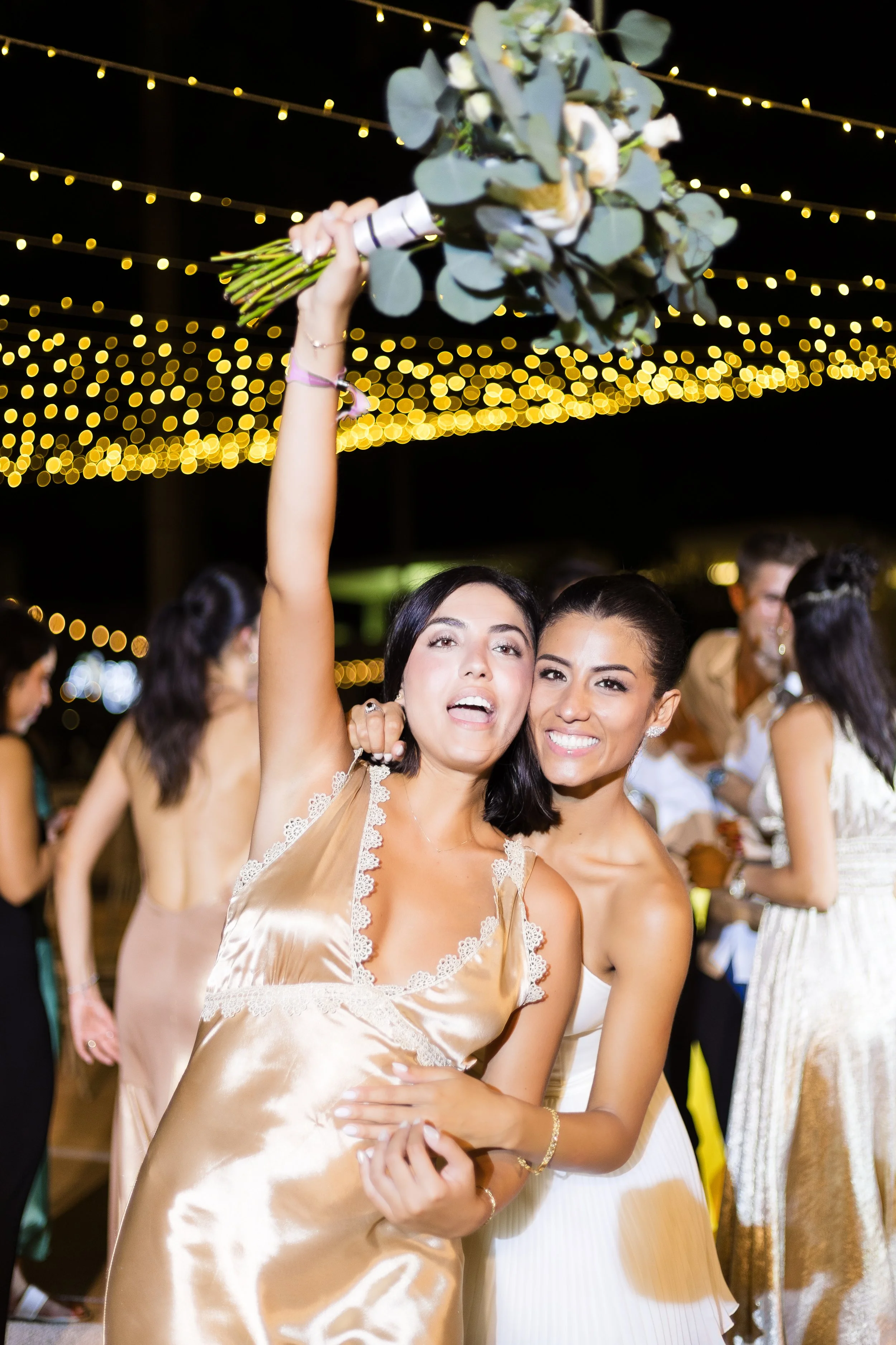 Two women smiling at a celebration, one holding a bouquet of eucalyptus and flowers above her head, with string lights in the background.