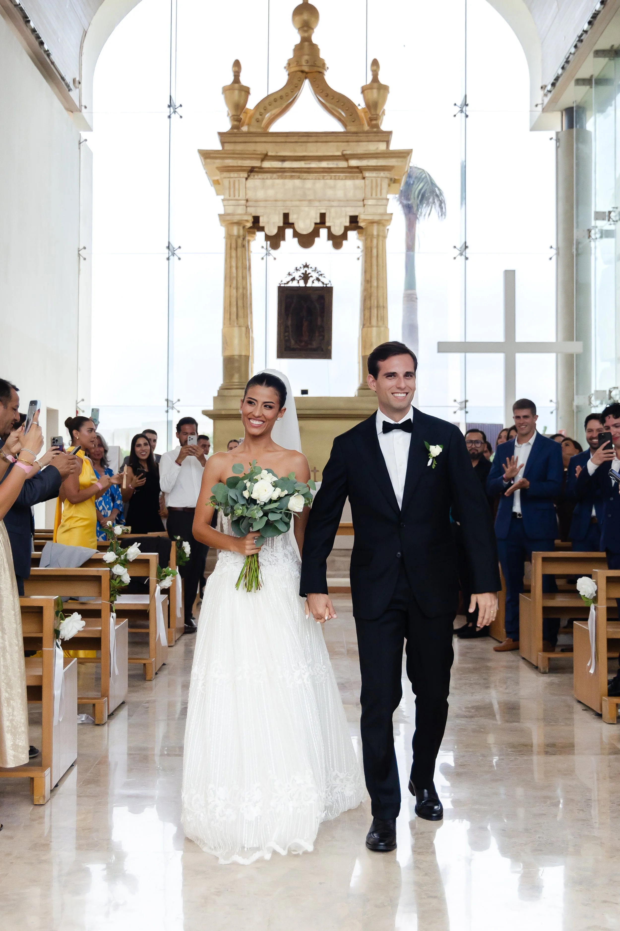 A bride and groom walking down the aisle in a church, holding hands, smiling, with guests taking photos and clapping in the background.