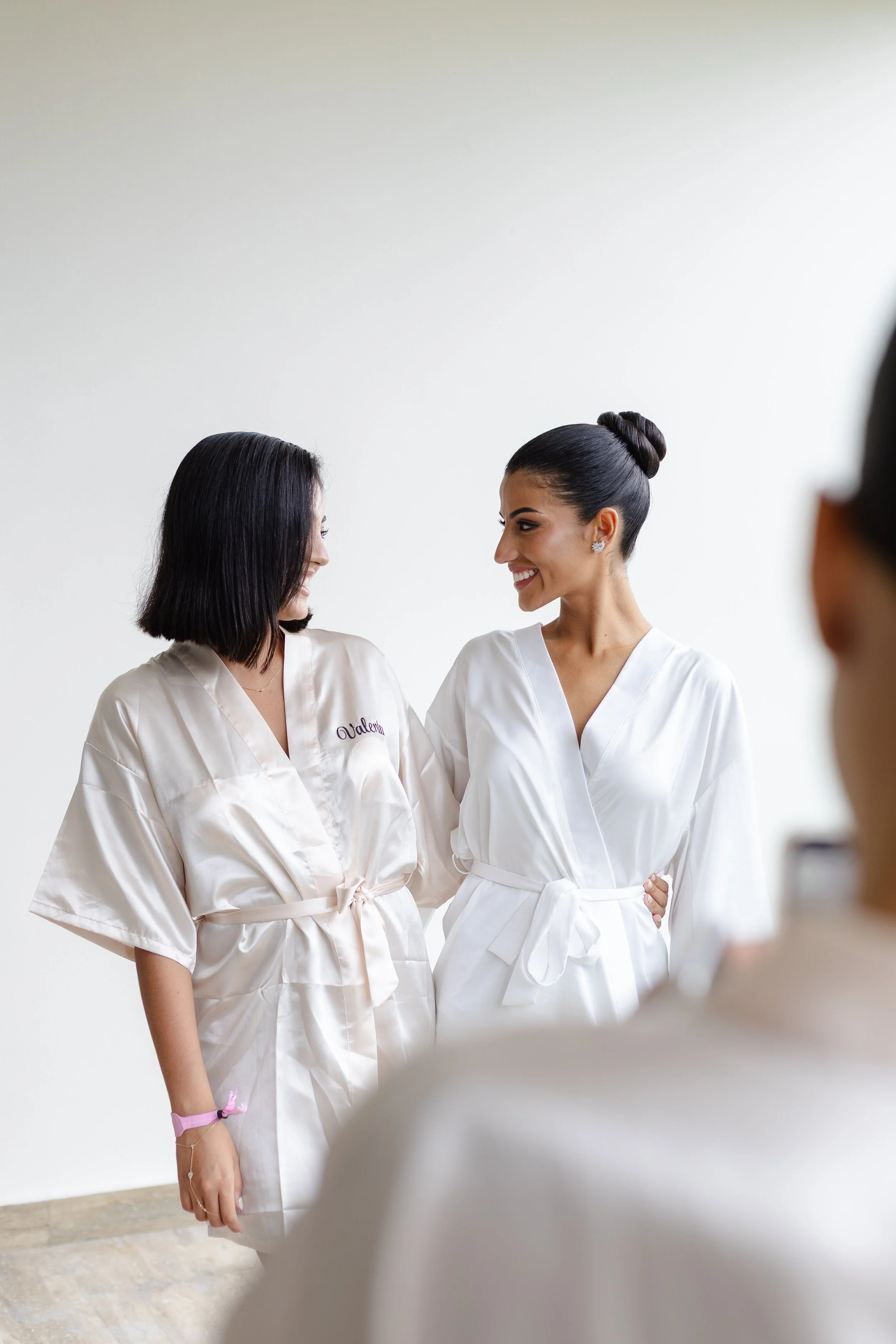 Two women in white robes smiling at each other, one with name embroidered 'Valeria', indoors with plain white wall background.