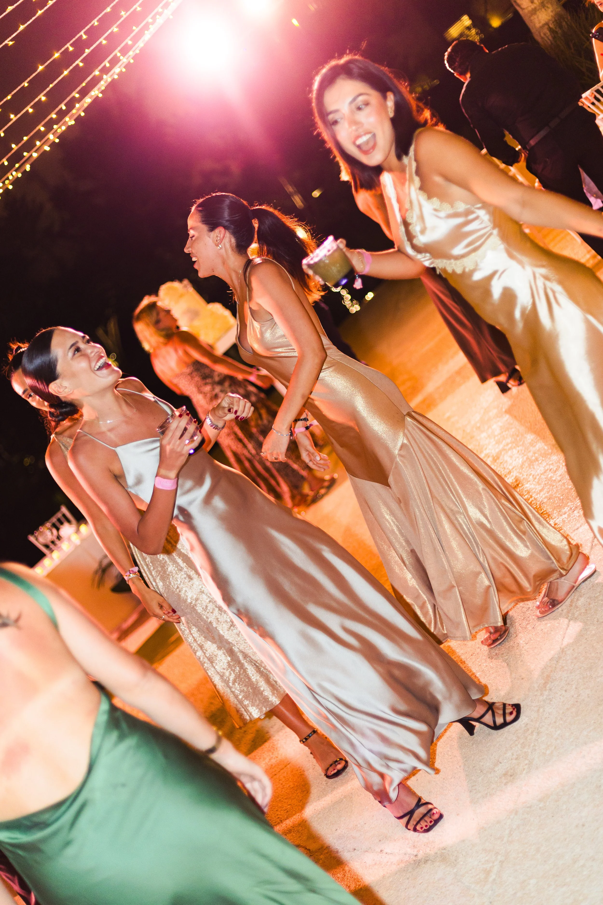 People in formal dresses dancing and celebrating at night with string lights overhead.
