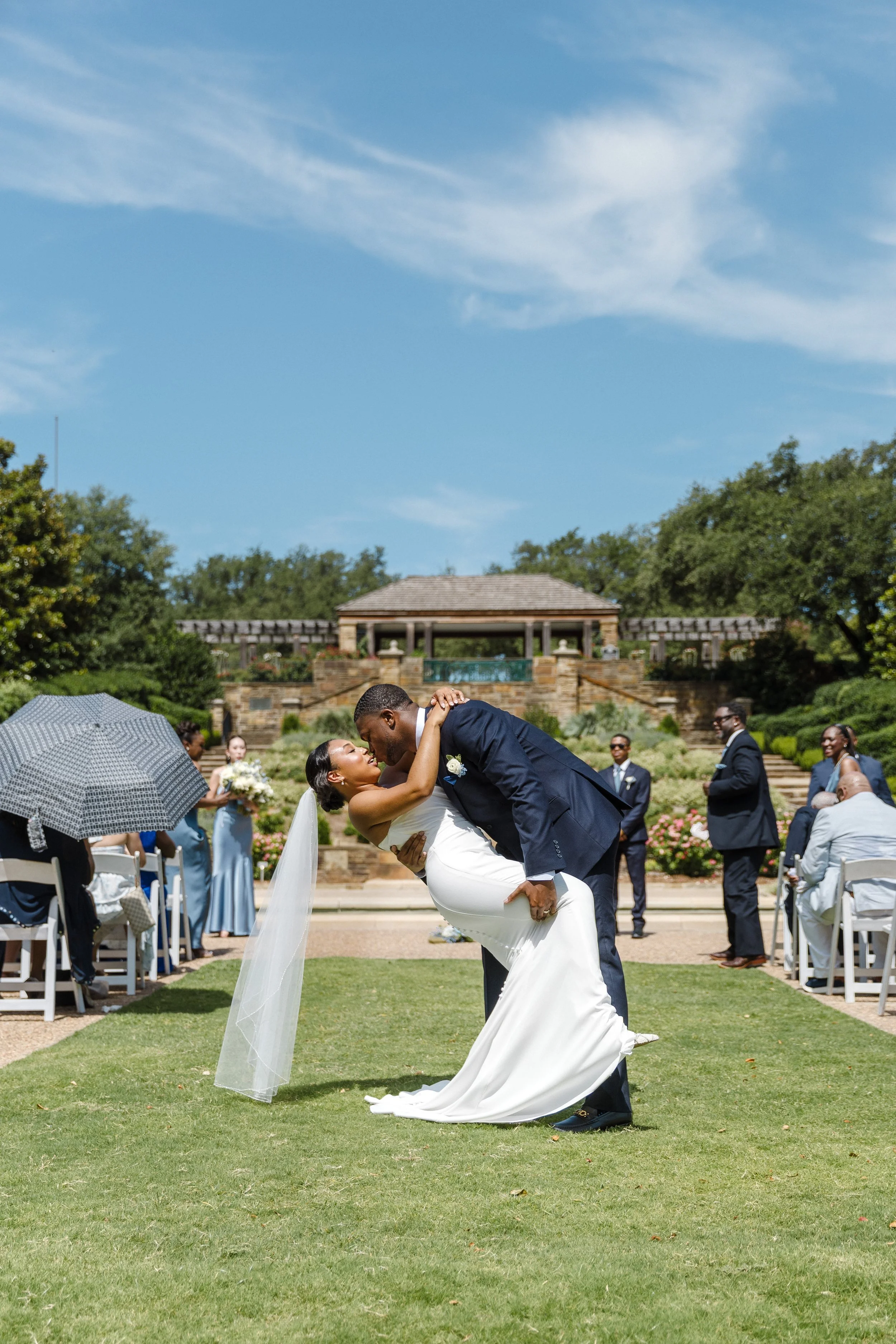 Couple sharing a kiss during their outdoor wedding ceremony, with guests seated on either side and a landscaped background under a blue sky.