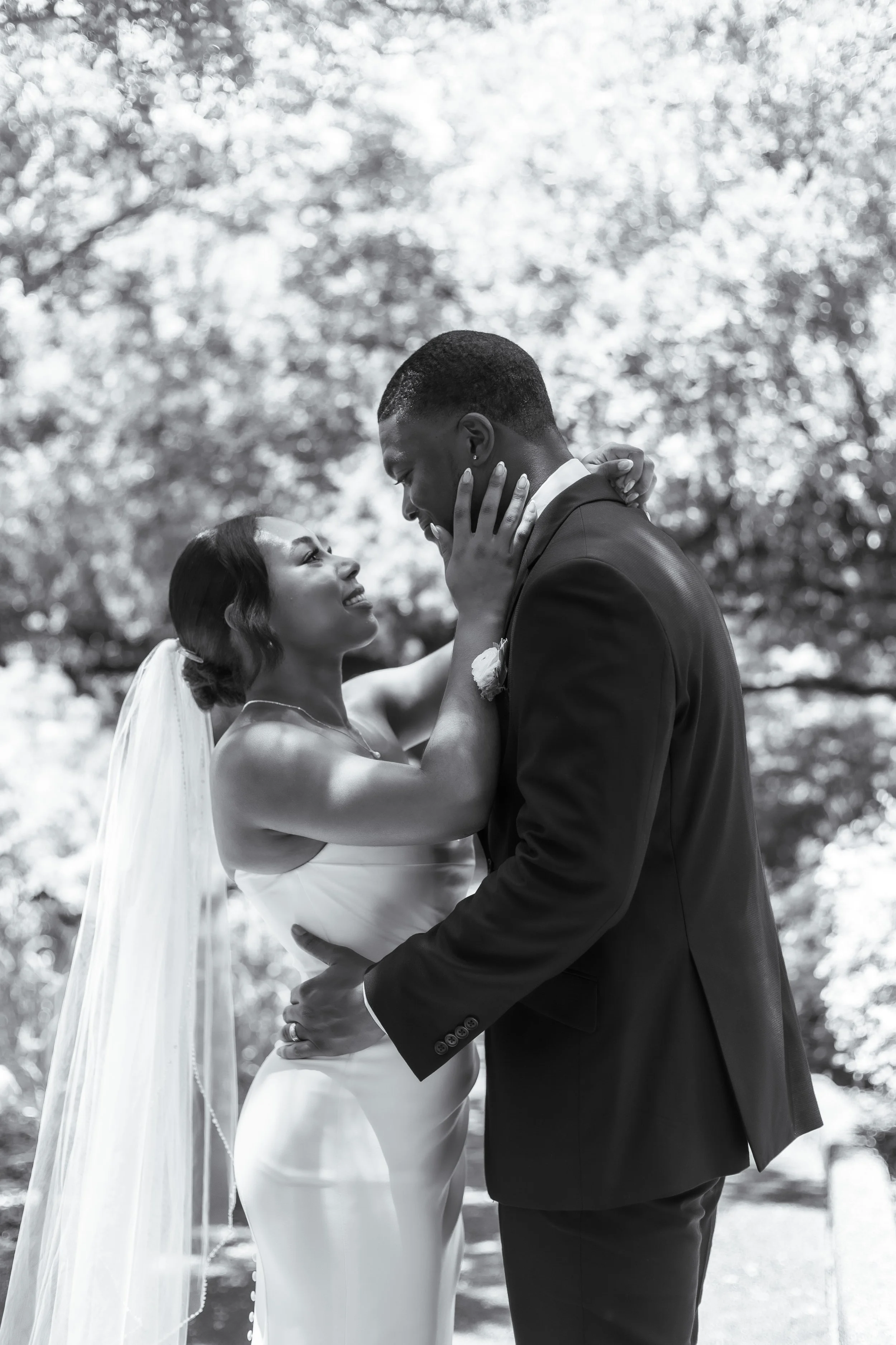 Black and white photo of a bride and groom gazing into each other's eyes, outdoors with trees in the background. The bride has wavy hair, a veil, and a sleeveless dress, while the groom is in a suit.