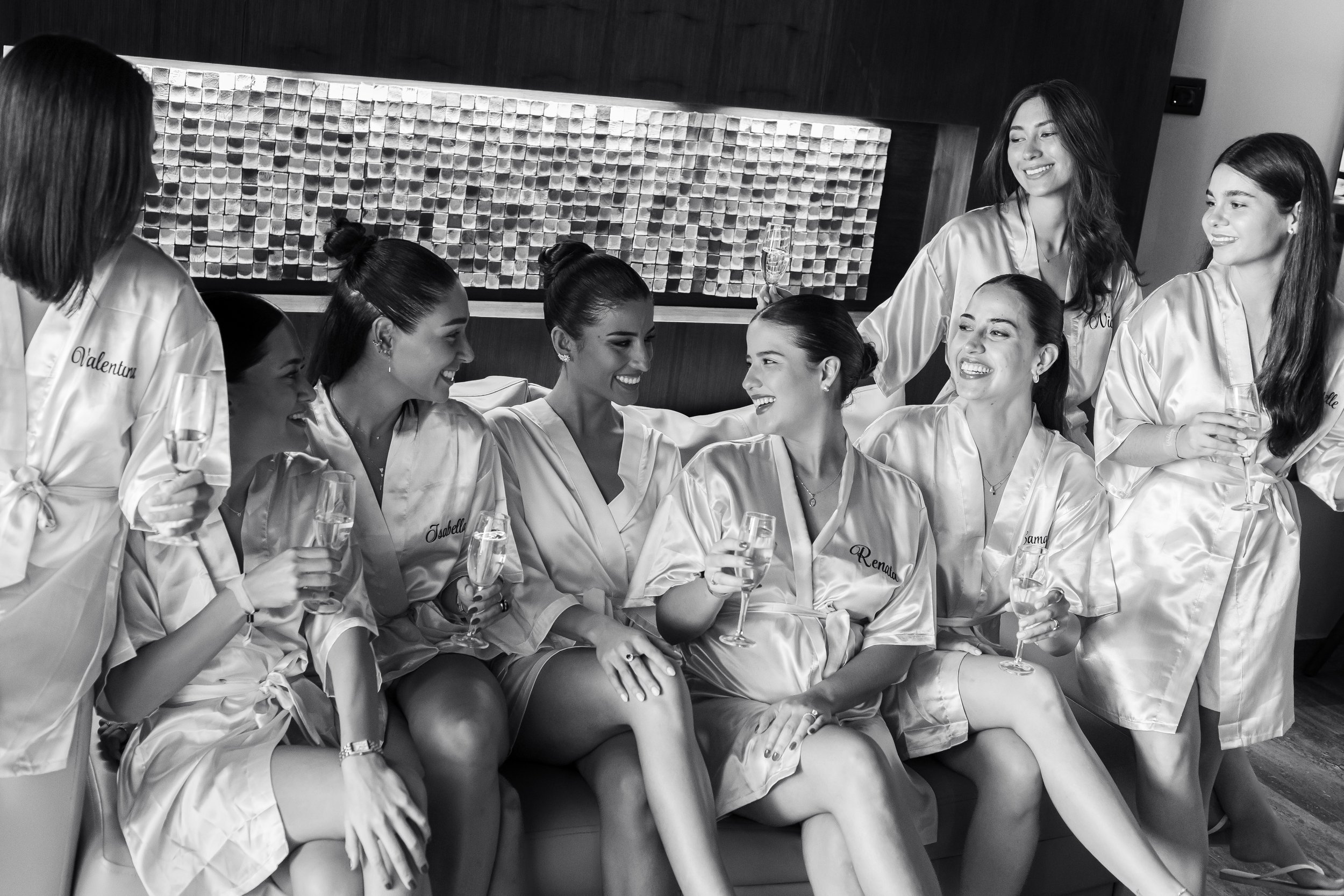 Group of women in matching satin robes, some holding champagne glasses, smiling and talking in a celebratory setting.