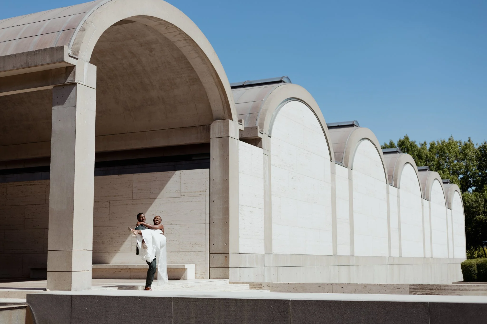 A couple dressed in wedding attire standing in front of a modern, white architectural building with large arches, under a clear blue sky.