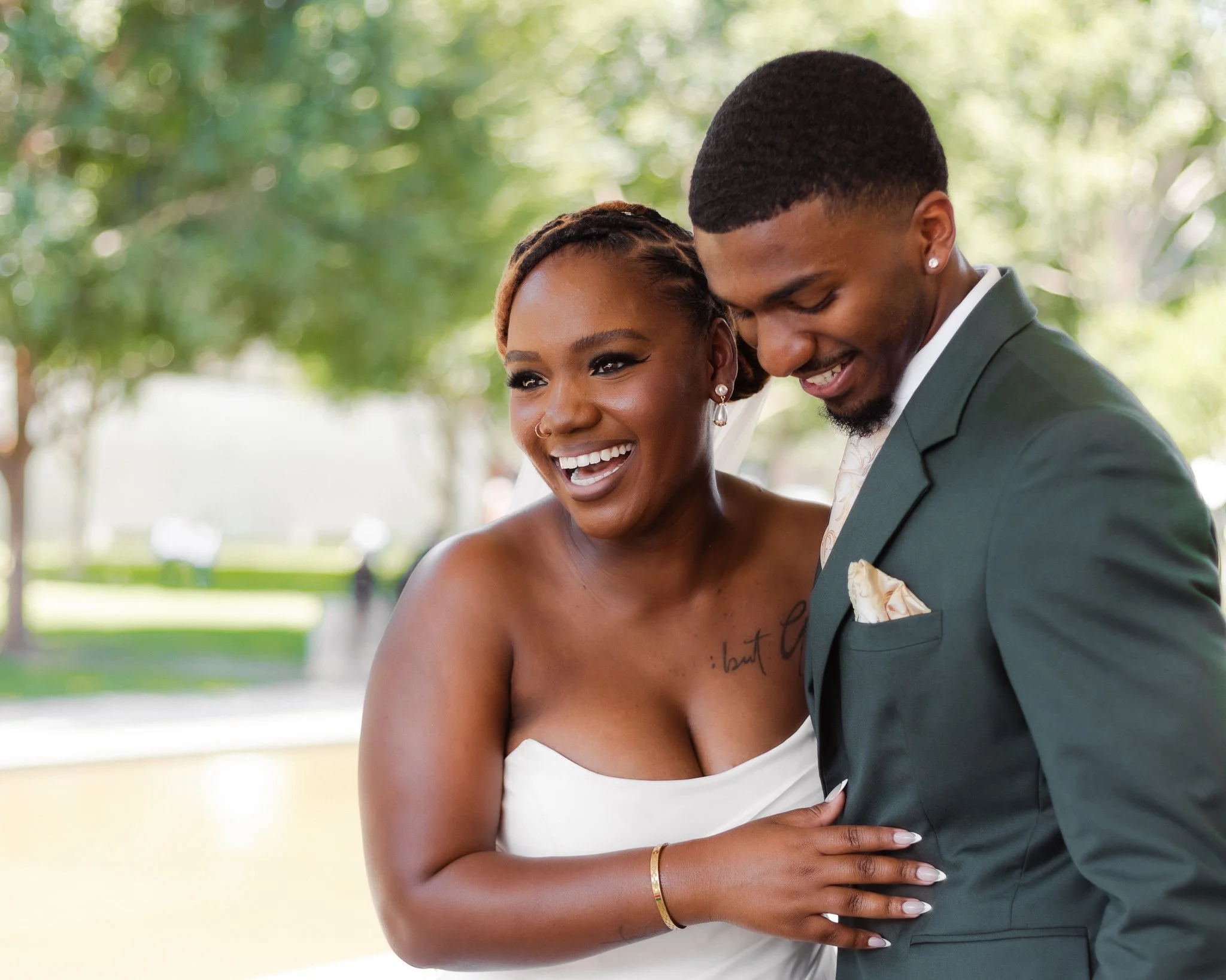 A smiling couple dressed in formal attire, sharing an intimate moment outdoors with lush green trees in the background.
