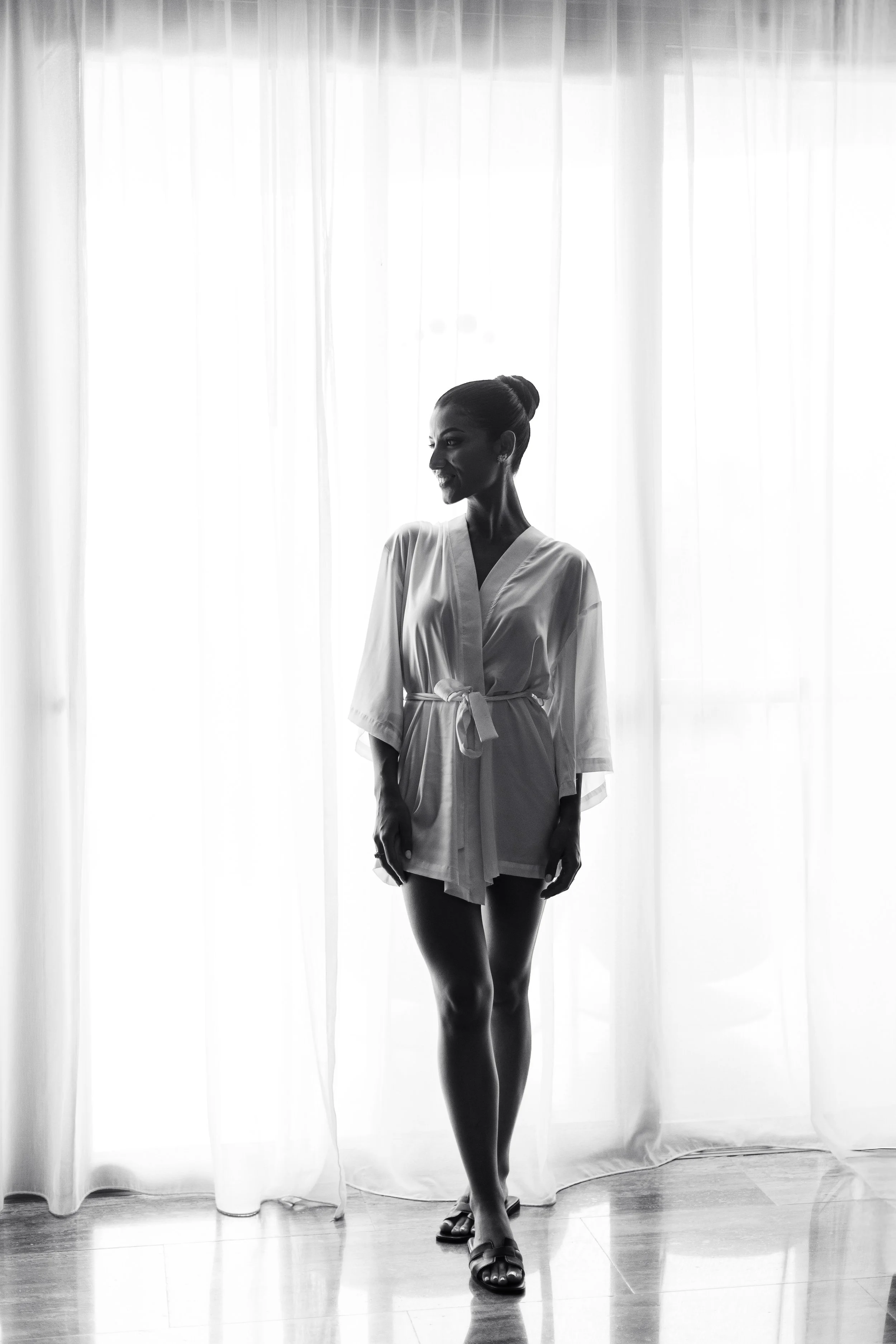 A woman in a white robe standing in front of a backlit curtain, smiling and looking to her left.