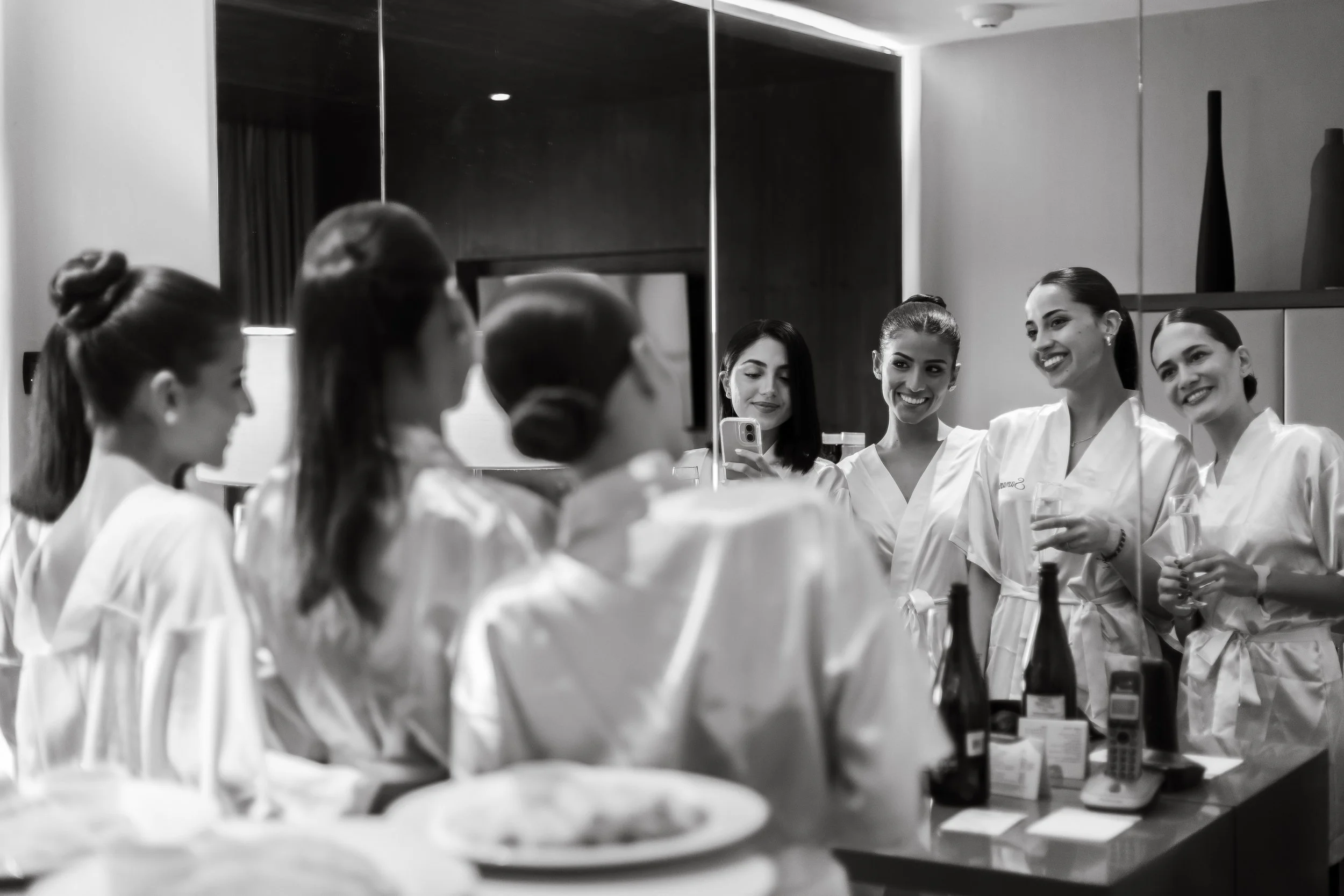 Group of women in white uniforms gathered around a mirror, smiling and chatting, some holding glasses, in a well-lit room with bottles on the table.