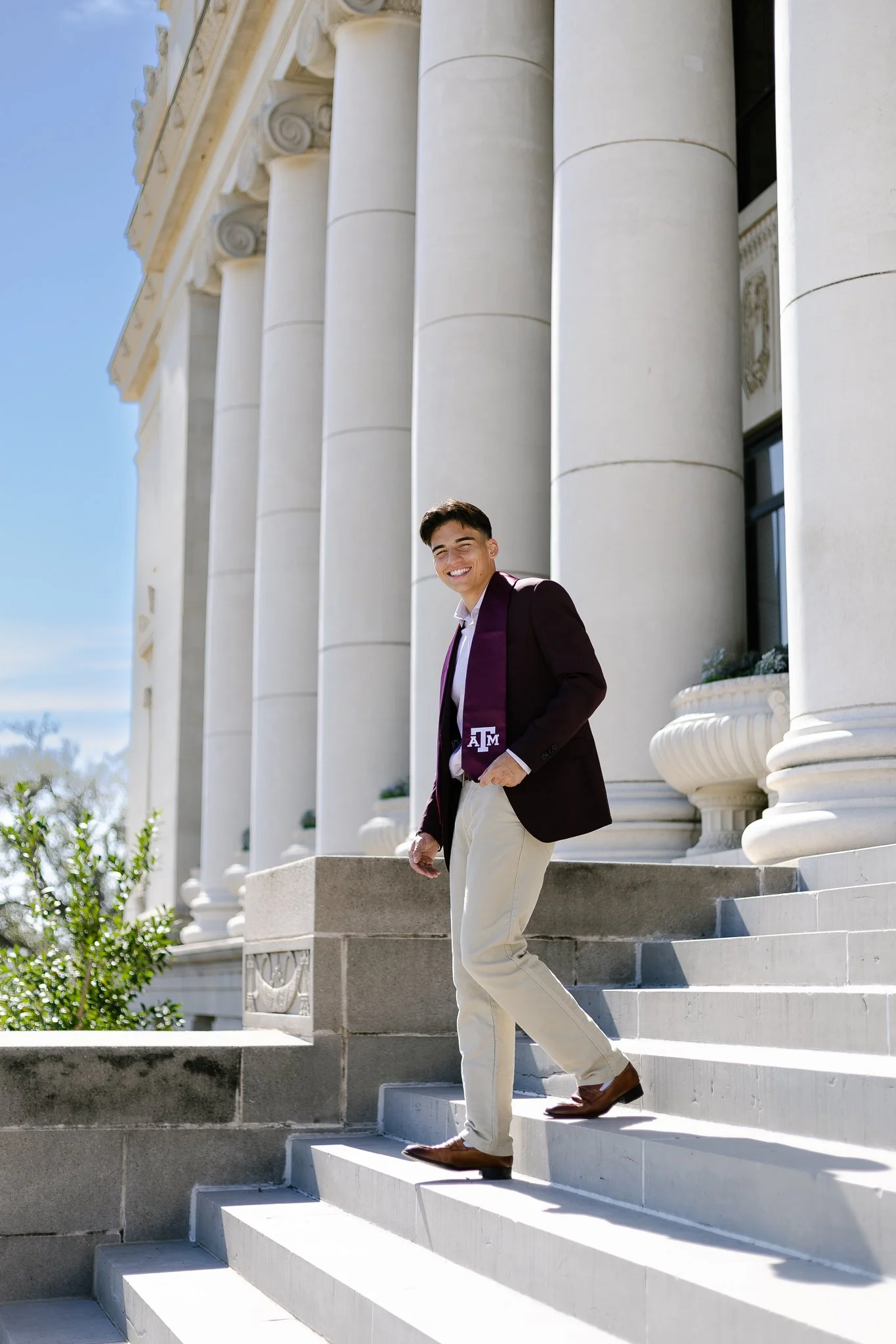 A young man in a maroon blazer, white shirt, and beige pants walking down steps outside a classical building with tall columns, smiling at the camera.