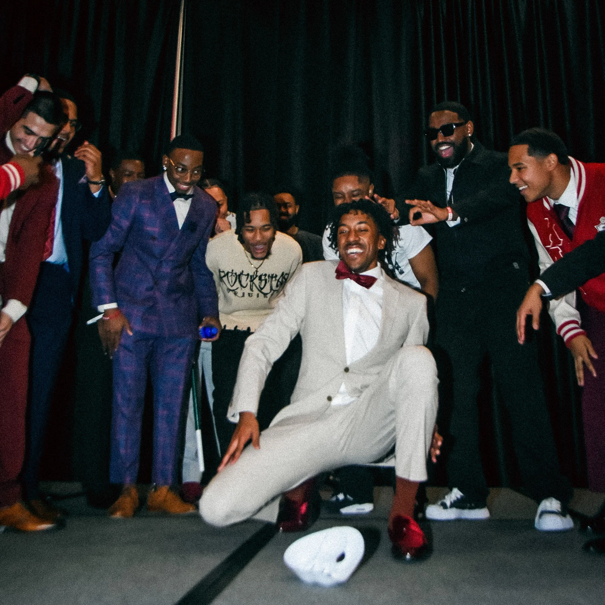 A group of men in formal attire, some with suits and bow ties, are gathered around a man in a white suit who is seated and smiling, while others are laughing and joking around.