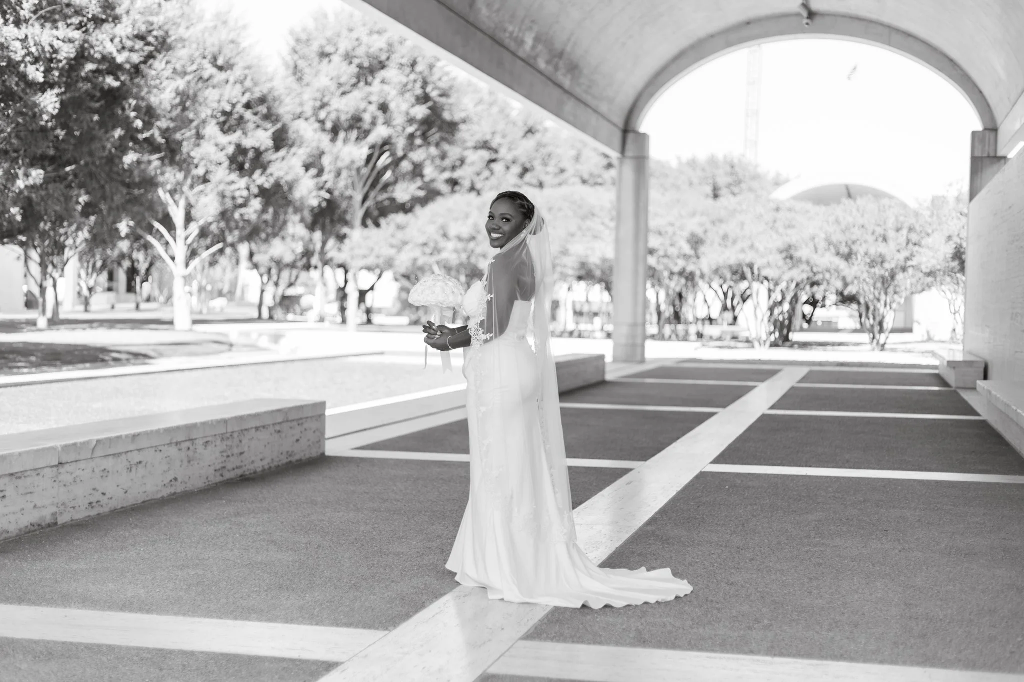 A bride in a wedding dress standing under an overpass, holding a bouquet, smiling at the camera with trees in the background.