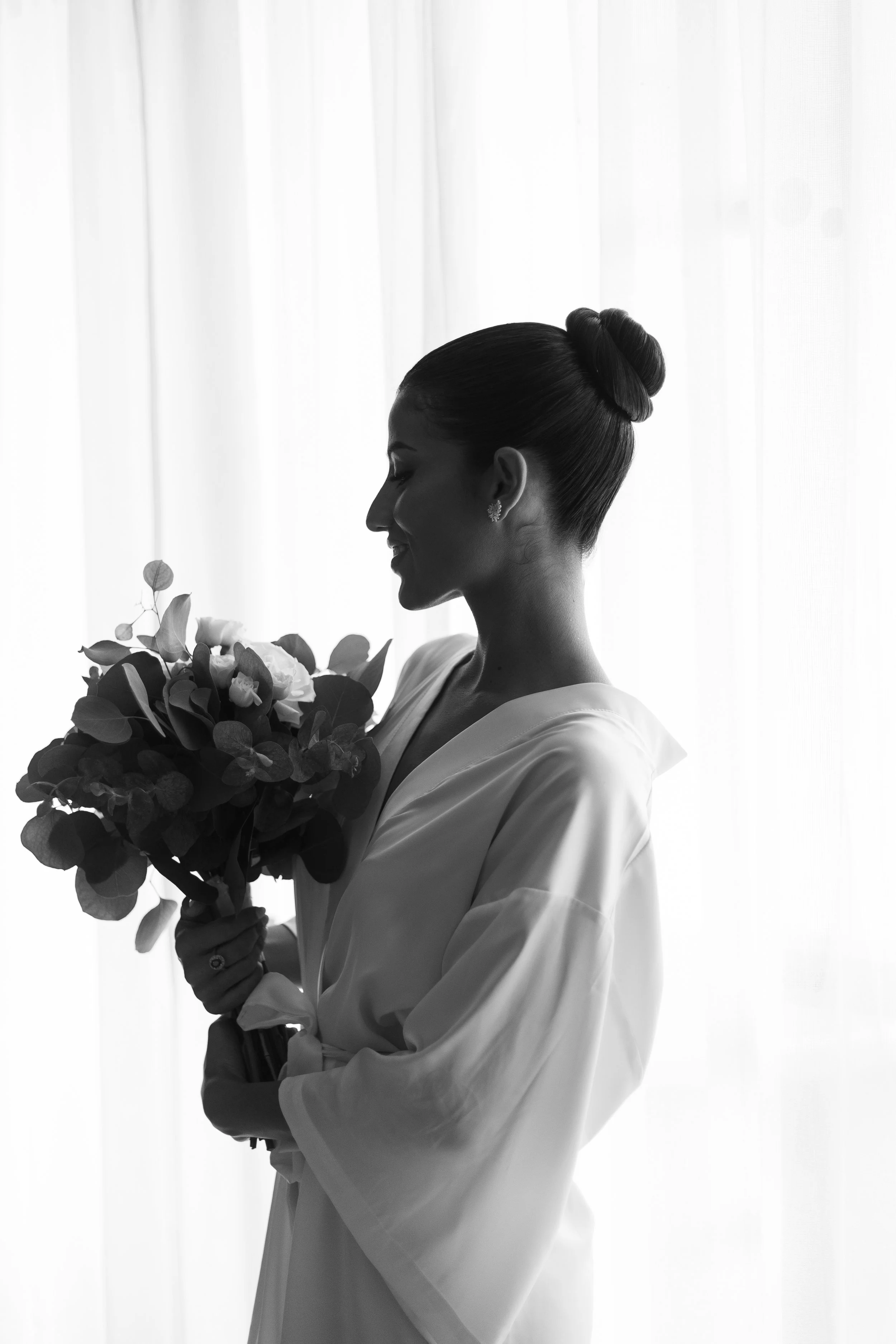 Silhouette of a woman in a robe holding a bouquet of flowers in front of a bright curtain.