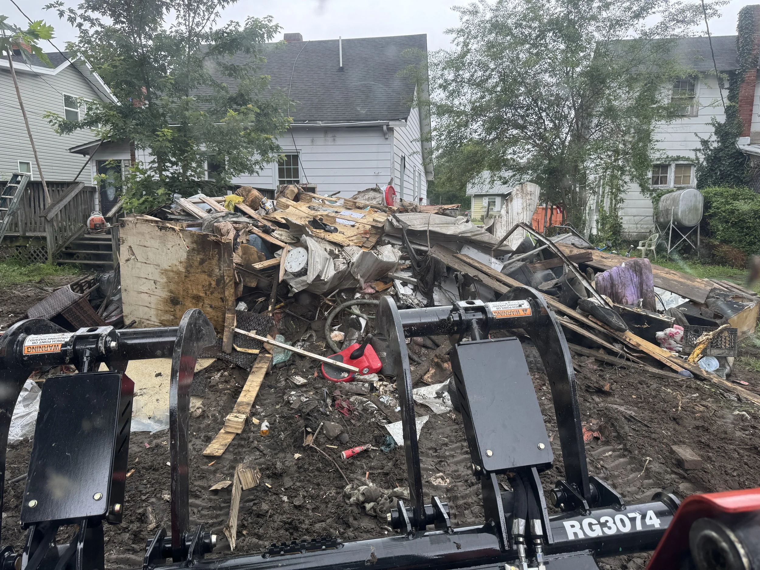 View of a destroyed house with scattered debris and mud, possibly after a natural disaster, with construction machinery in the foreground.