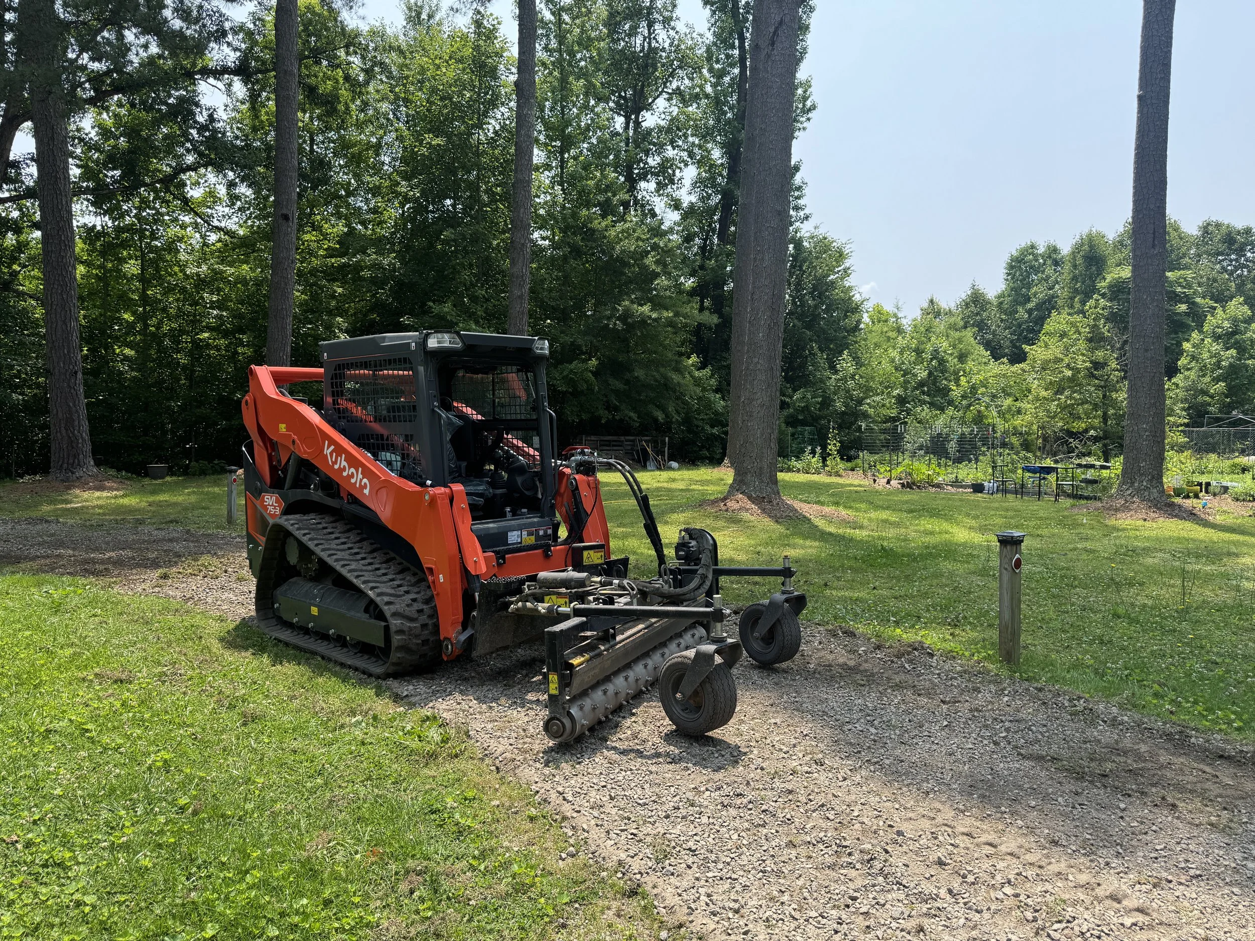 A Kubota compact tracked utility vehicle parked on a gravel path in a grassy area with trees in the background.