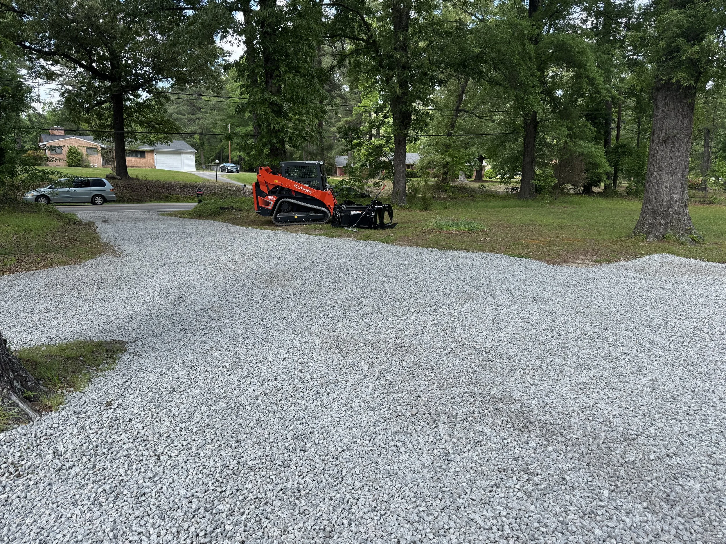Gravel driveway with a red compact track loader and attachments, surrounded by trees and a residential neighborhood in the background.