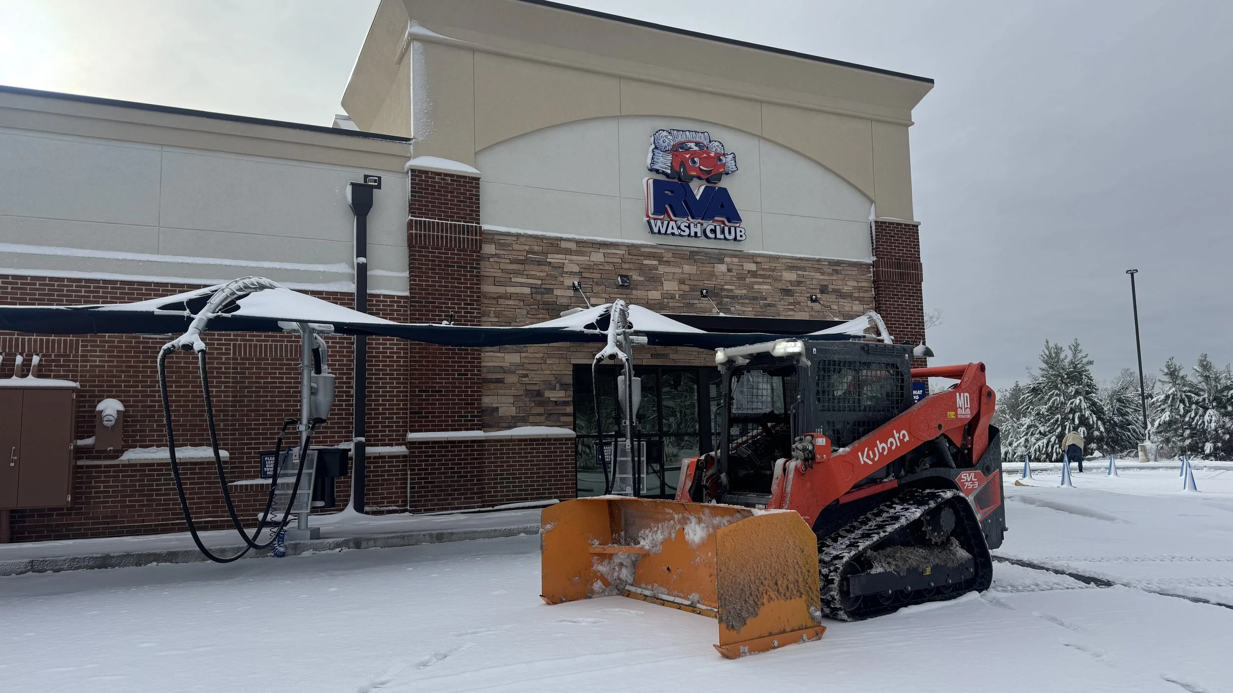 Snow removal equipment in front of RVA Wash Club, a brick and stucco building with a sign featuring a cartoon car. The ground and surroundings are covered in snow, and a person is seen in the distance near snow-covered trees.