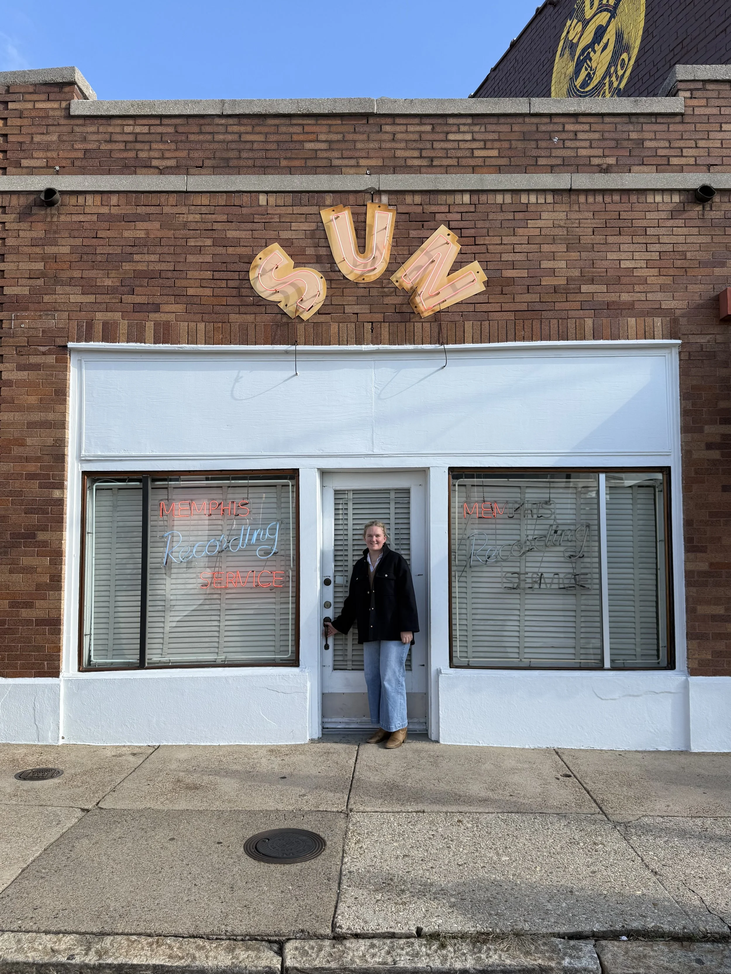 Woman standing in front of a brick storefront with neon signs reading Memphis Recording Service and Sul Neon sign on top of the building.