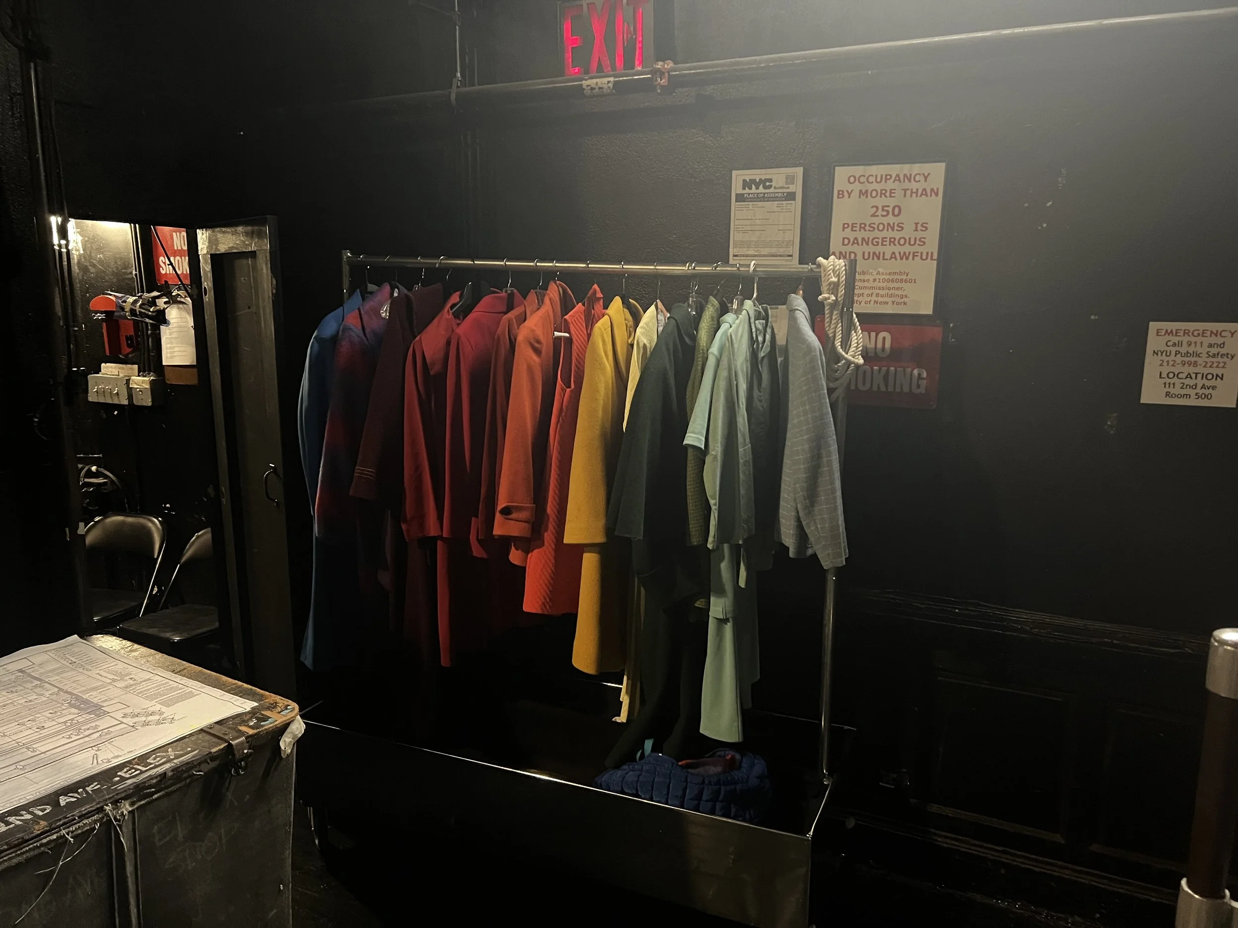 Clothing rack with colorful jackets and shirts in a dimly lit backstage area, with a mirror, chair, and posters on the wall.
