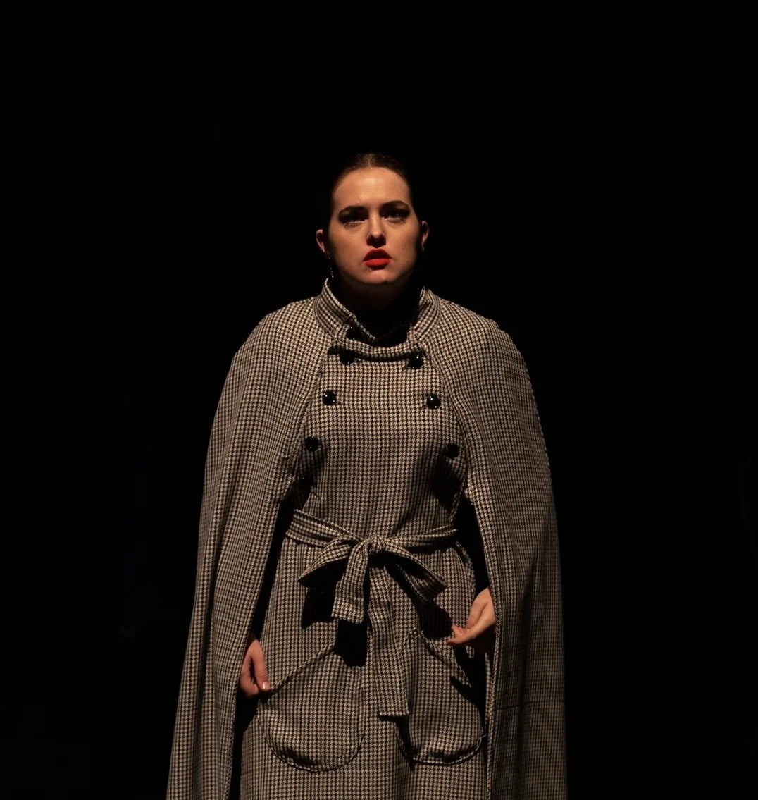 A woman standing in a spotlight against a dark background, wearing a checkered coat with a tied belt and a cape, with neutral makeup and red lipstick.