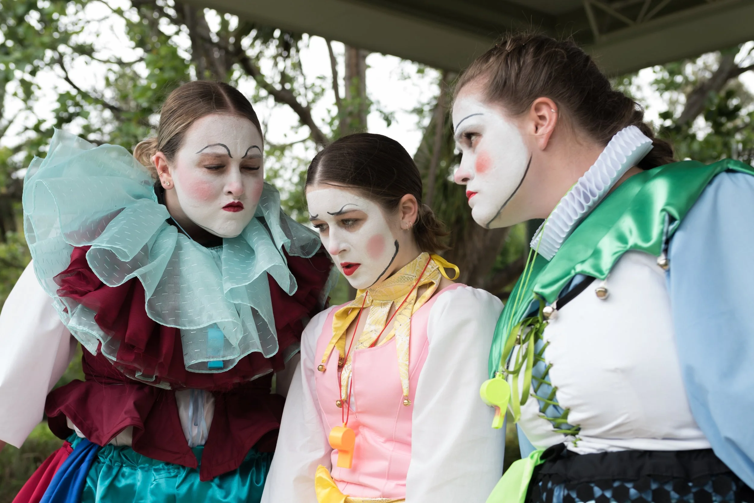 Three performers dressed as clowns with white face paint, colorful costumes, and exaggerated expressions, looking downward during an outdoor performance.
