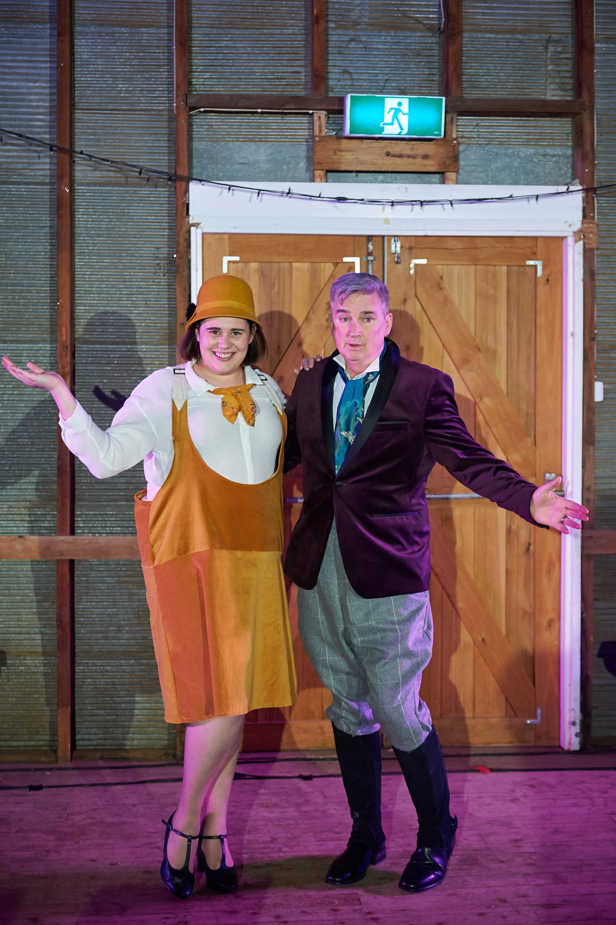 Two people dressed in theatrical costumes standing in front of wooden doors under an emergency exit sign, both smiling with arms outstretched.