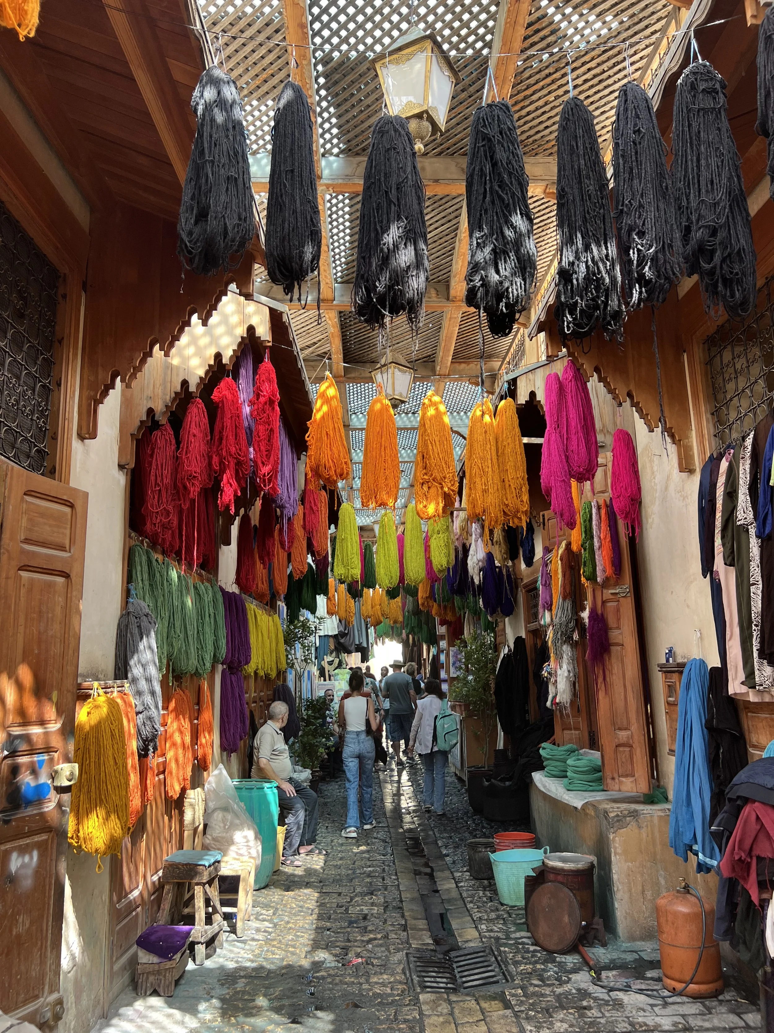 Colorful yarns displayed in a marketplace with people walking and shopping.