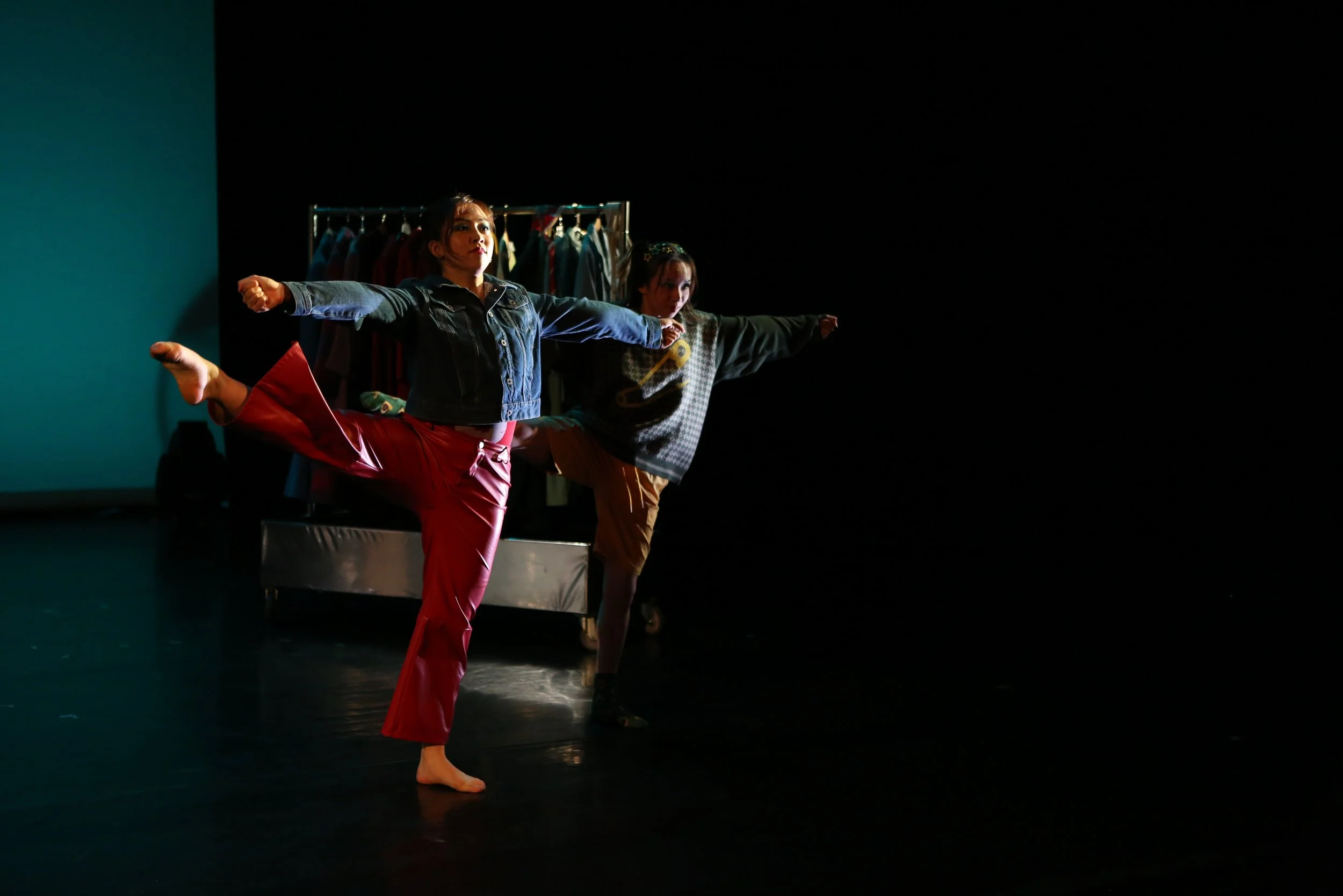 Two women practicing dance or yoga on stage with clothing rack in the background, one woman in red pants and denim jacket lifting her leg, the other in brown pants and hoodie.