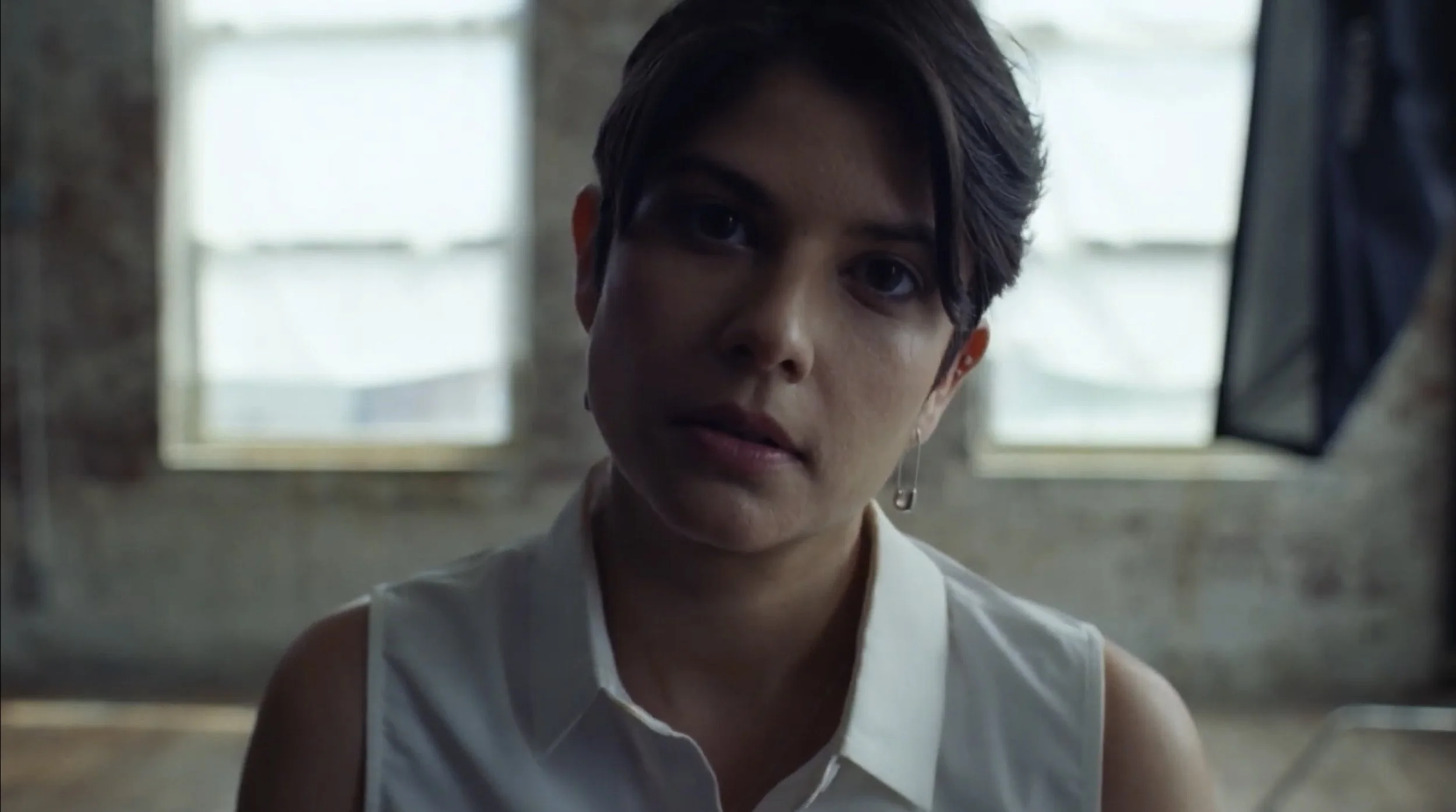Close-up of a woman with short dark hair and earrings, wearing a white sleeveless top, looking at the camera with a serious expression inside a room with concrete walls and windows in the background.