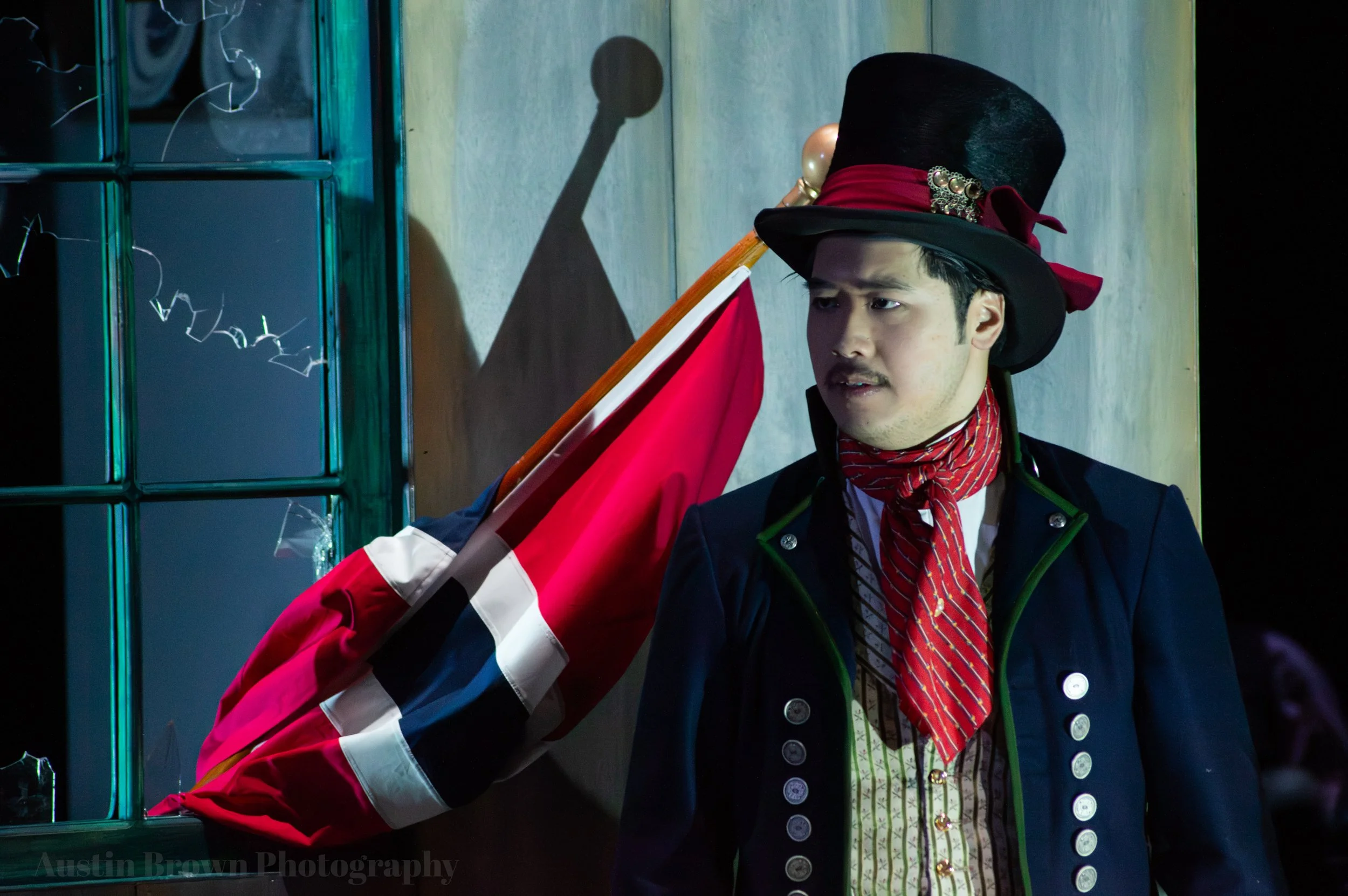 A man dressed in historical costume, wearing a black top hat with a red ribbon and a bow, a navy blue coat with buttons, a red scarf, and holding a flag with red, white, blue, and black colors, stands against a wall with a broken window. A shadow of a baseball bat is cast on the wall behind him.