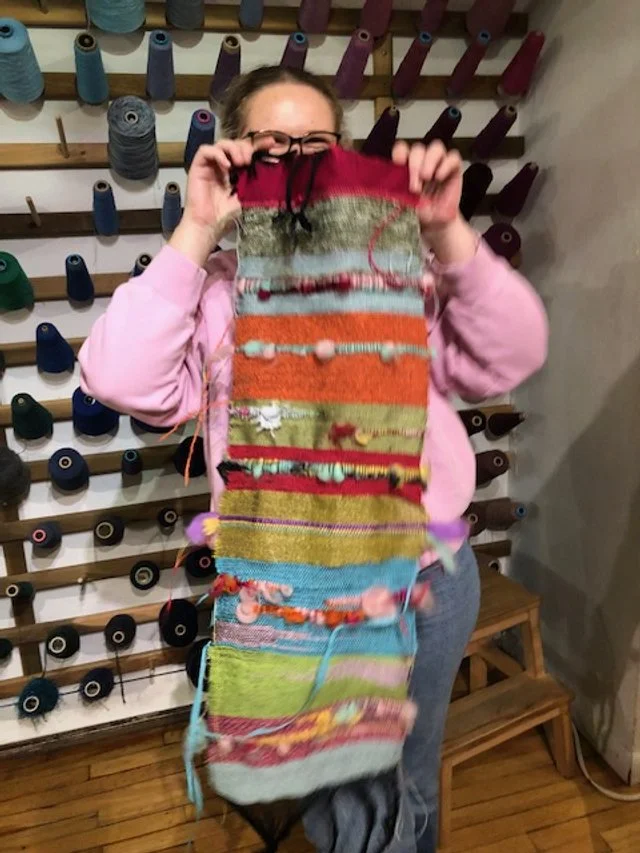 Person holding a colorful knitted or woven piece of fabric with various textures and colors, behind spools of thread on a wall.