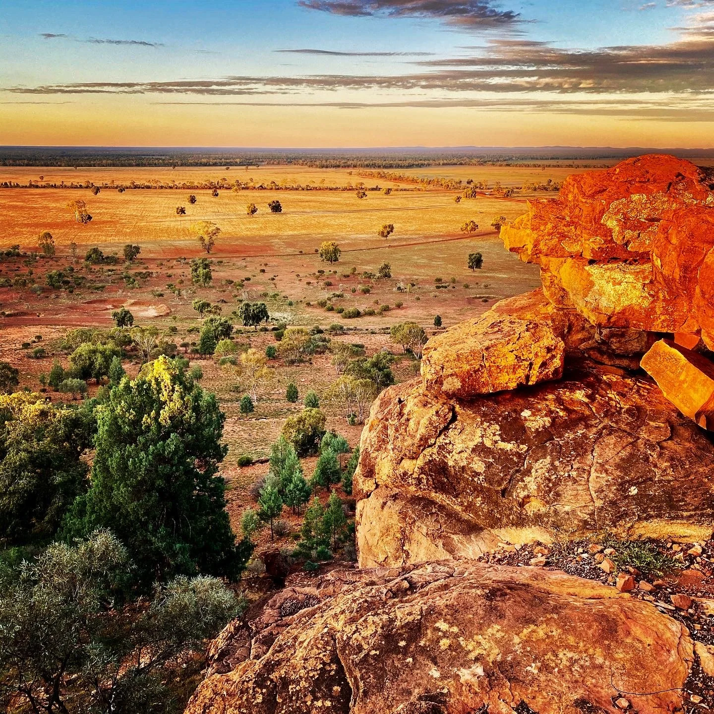 Scenic view of an arid landscape during sunset, featuring large orange rocks in the foreground, scattered trees, and a vast horizon with a colorful sky.