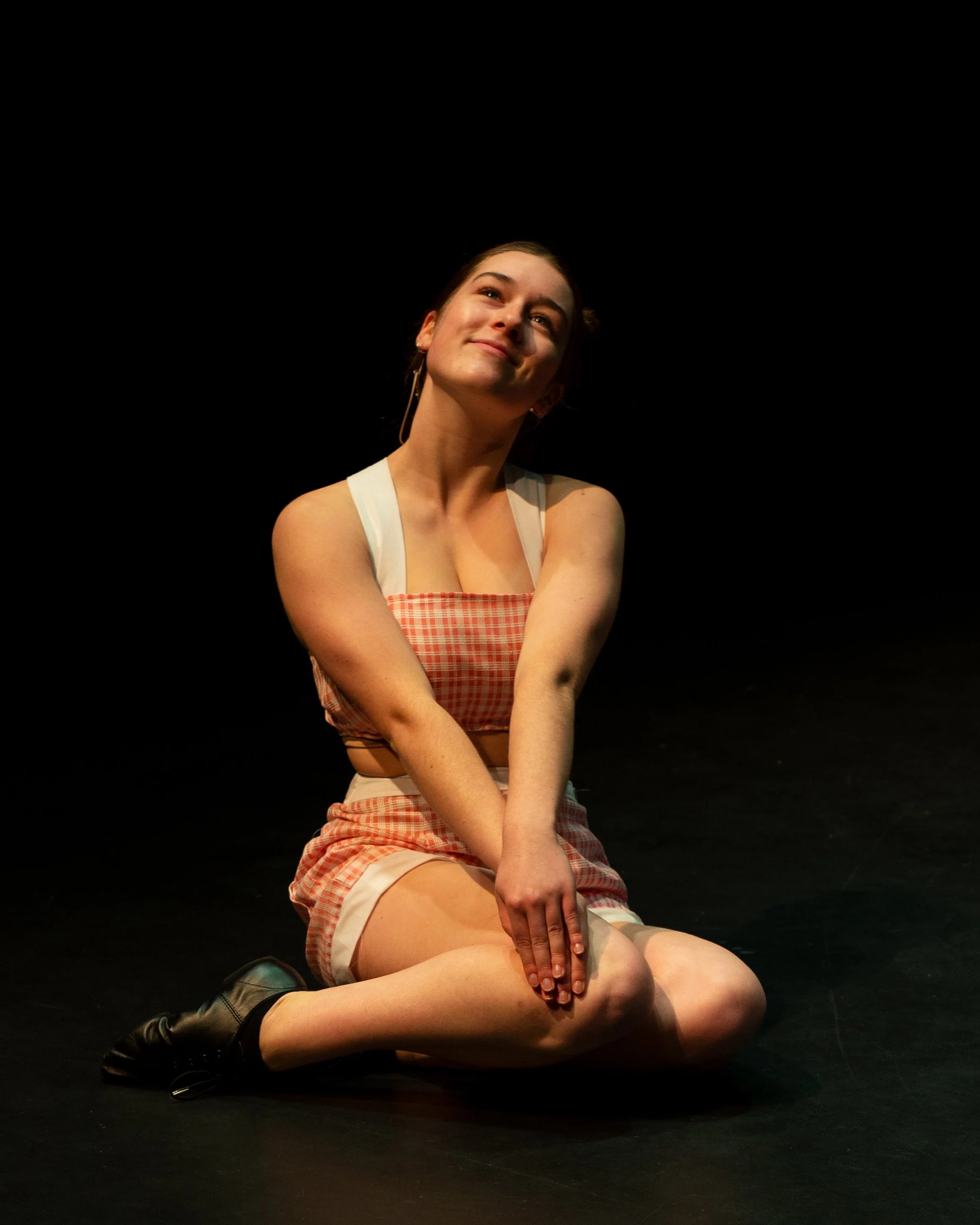 A young woman with brown hair, wearing a checkered crop top and matching shorts, is kneeling on the floor with her arms crossed over her knees, smiling and looking upwards, against a black background.