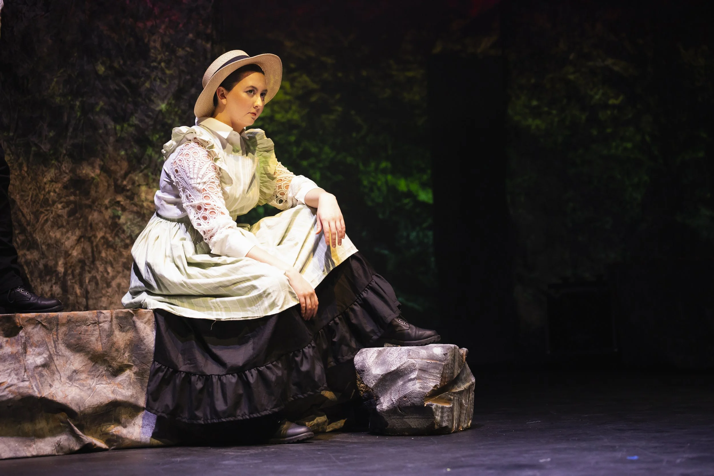 Woman dressed in period costume sitting on a rocky stage set, with a dark background and some green lighting.