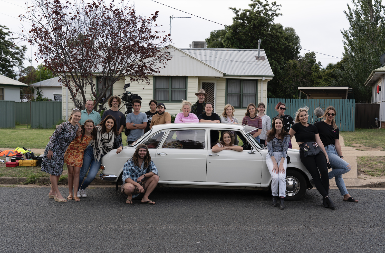 Group of people gathered around a white vintage car in front of a house, with some standing and some sitting, on a suburban street.