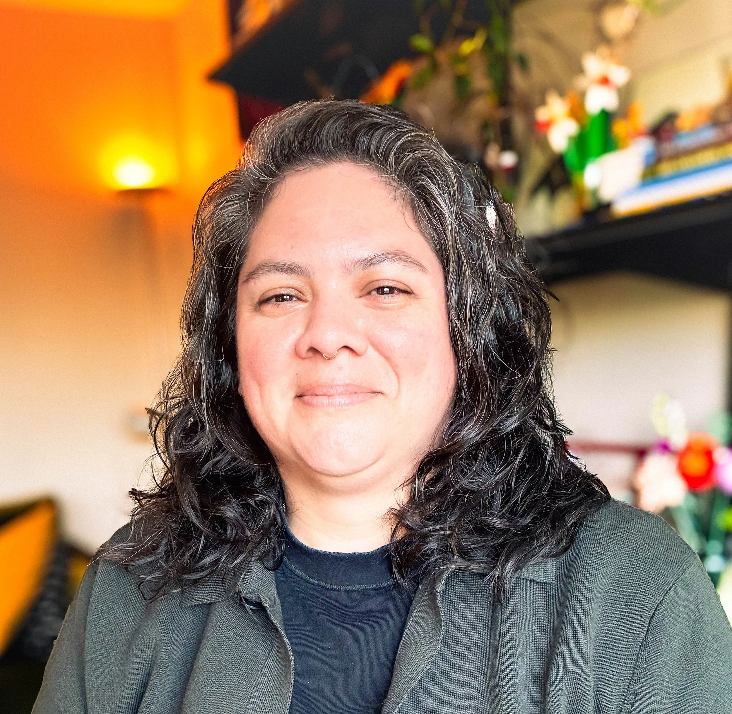 A non-binary person with shoulder length, dark brown way hair, wearing a septum ring and smiling. They're sitting indoors with warm lighting, decorative plants, and colorful items in the background.
