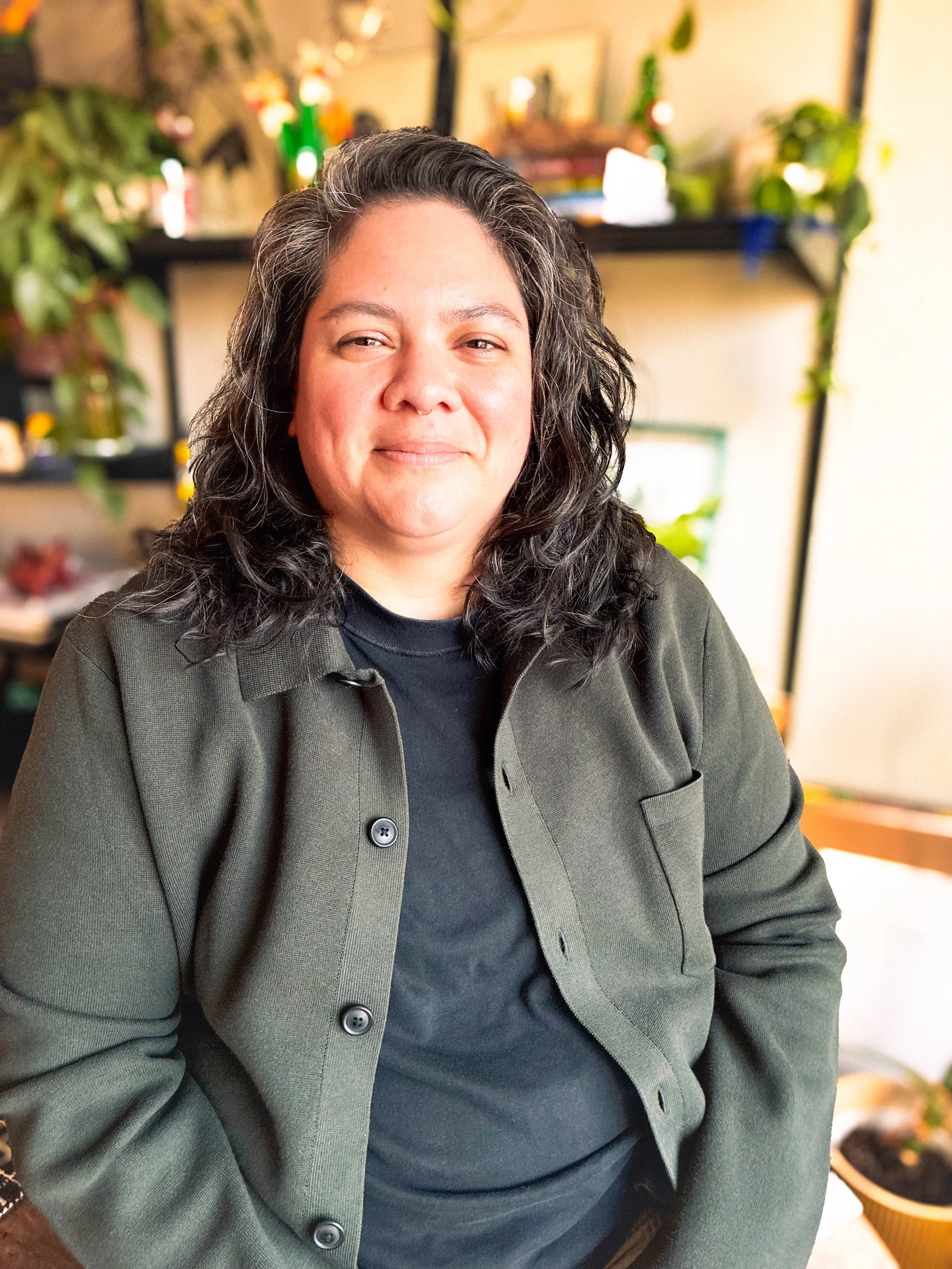 A non-binary person with shoulder length, dark brown wavy hair and light skin, wearing a dark green button-up over a black t-shirt, sitting indoors in a cozy, plant-filled room.