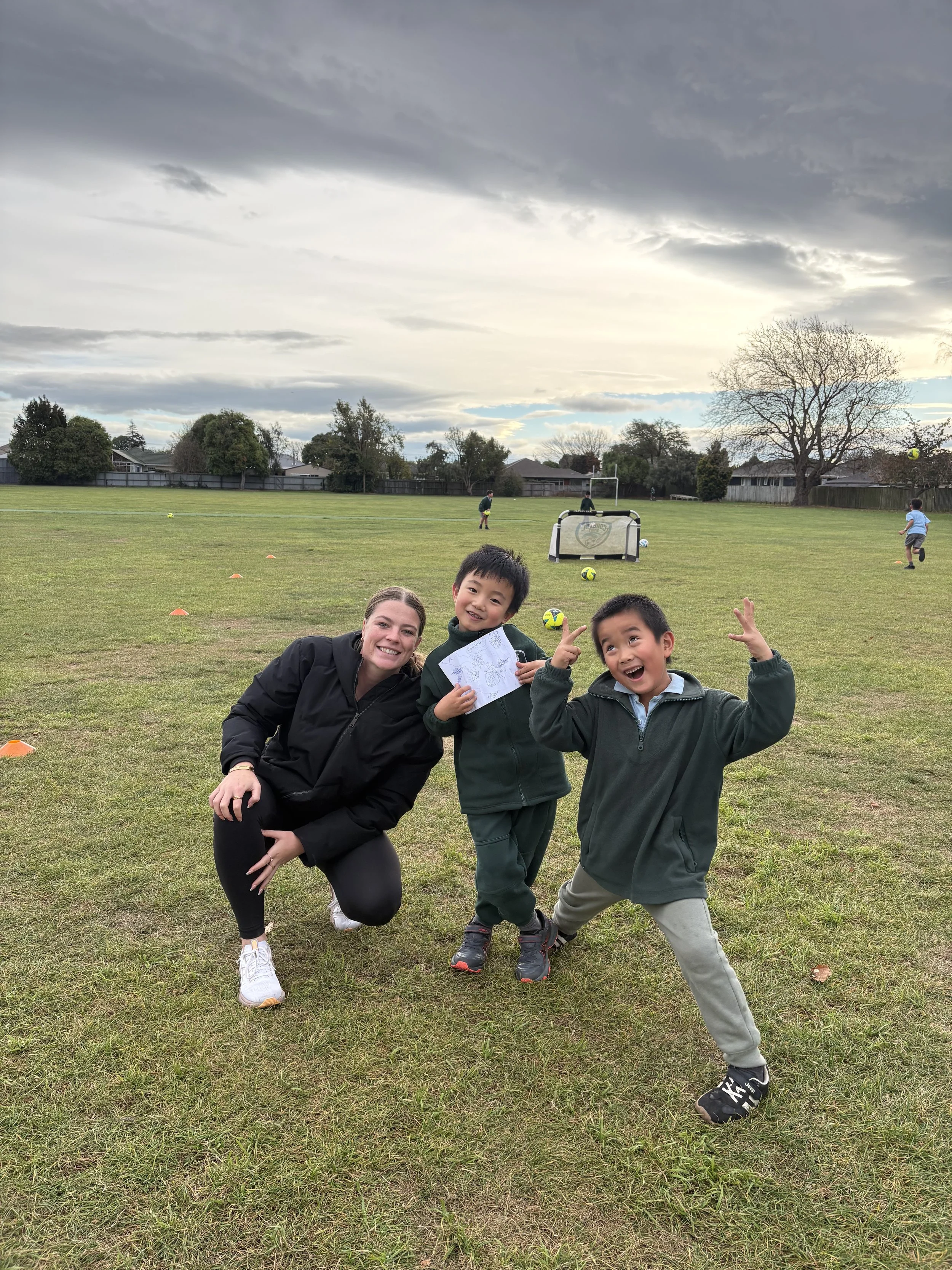Children and an adult on a grassy field, with a playground in the background and a cloudy sky overhead.