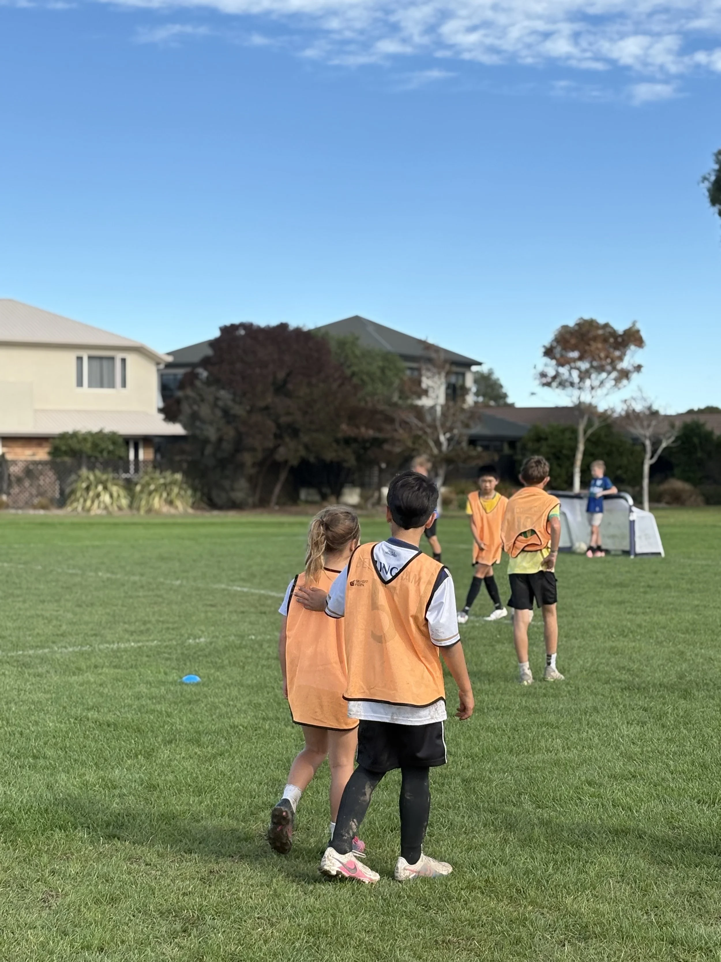 Children playing soccer on a grassy field with houses and trees in the background under a blue sky.