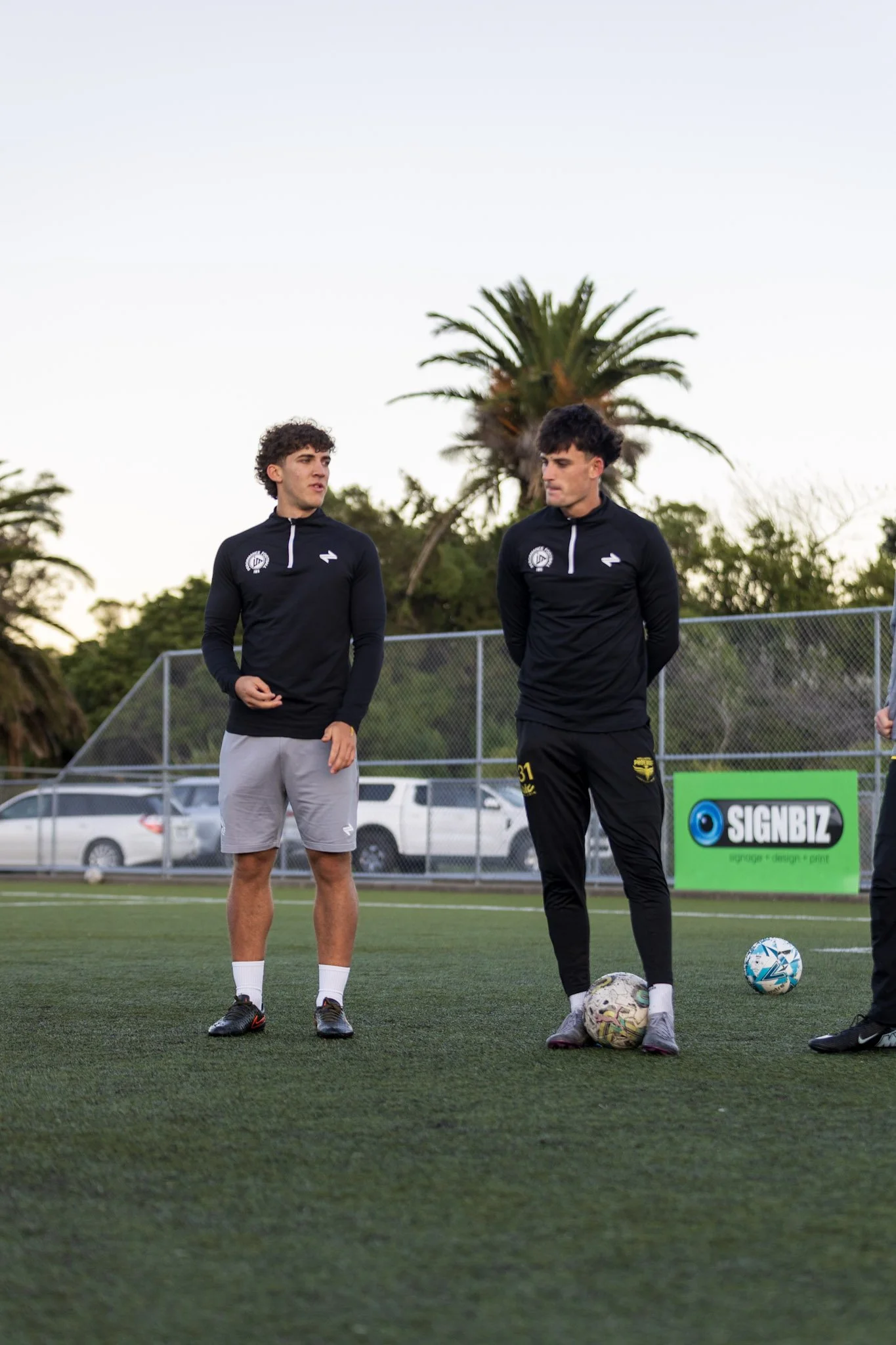 Young man in blue soccer jersey running on a soccer field with a soccer ball, with another person observing in the background.