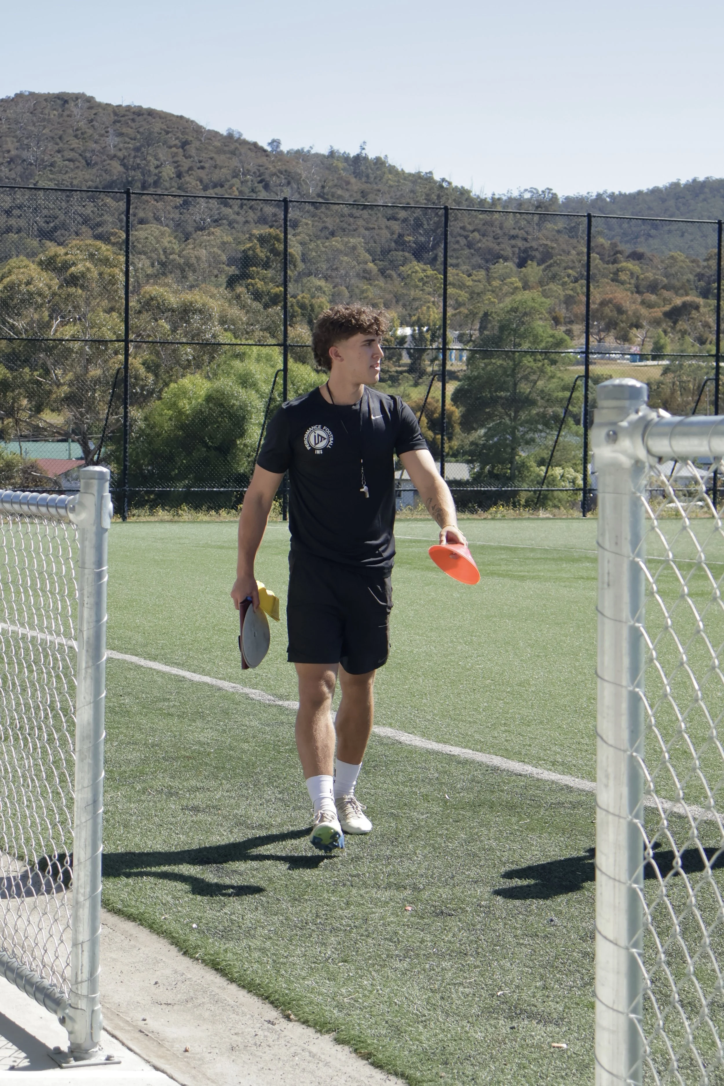 A young male soccer coach walking on the field holding a referee paddle in each hand, wearing a black shirt with a logo and black shorts, with a whistle around his neck.