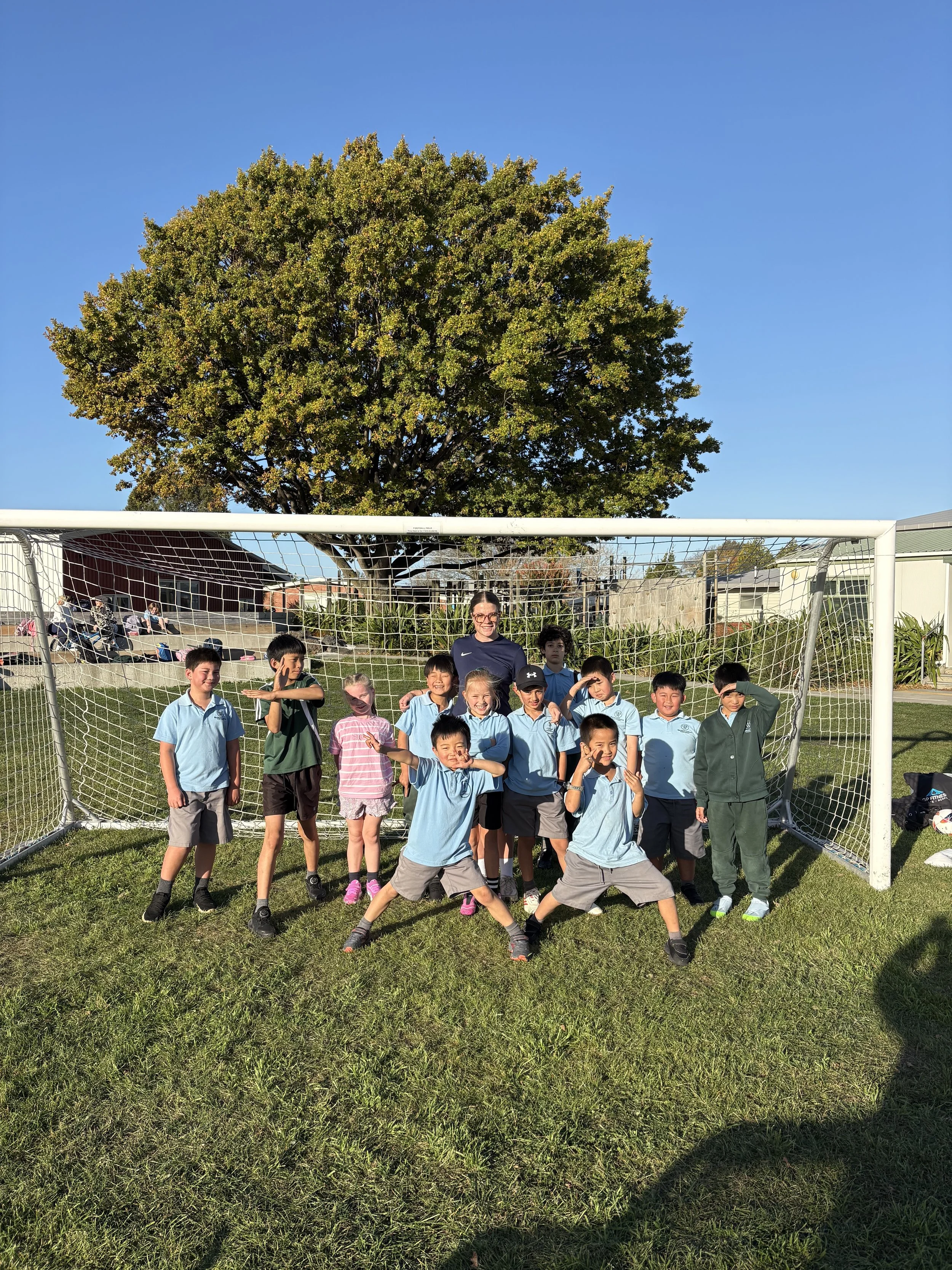 Group of children and a woman posing in front of a soccer goal on a grassy field under a large tree on a sunny day.