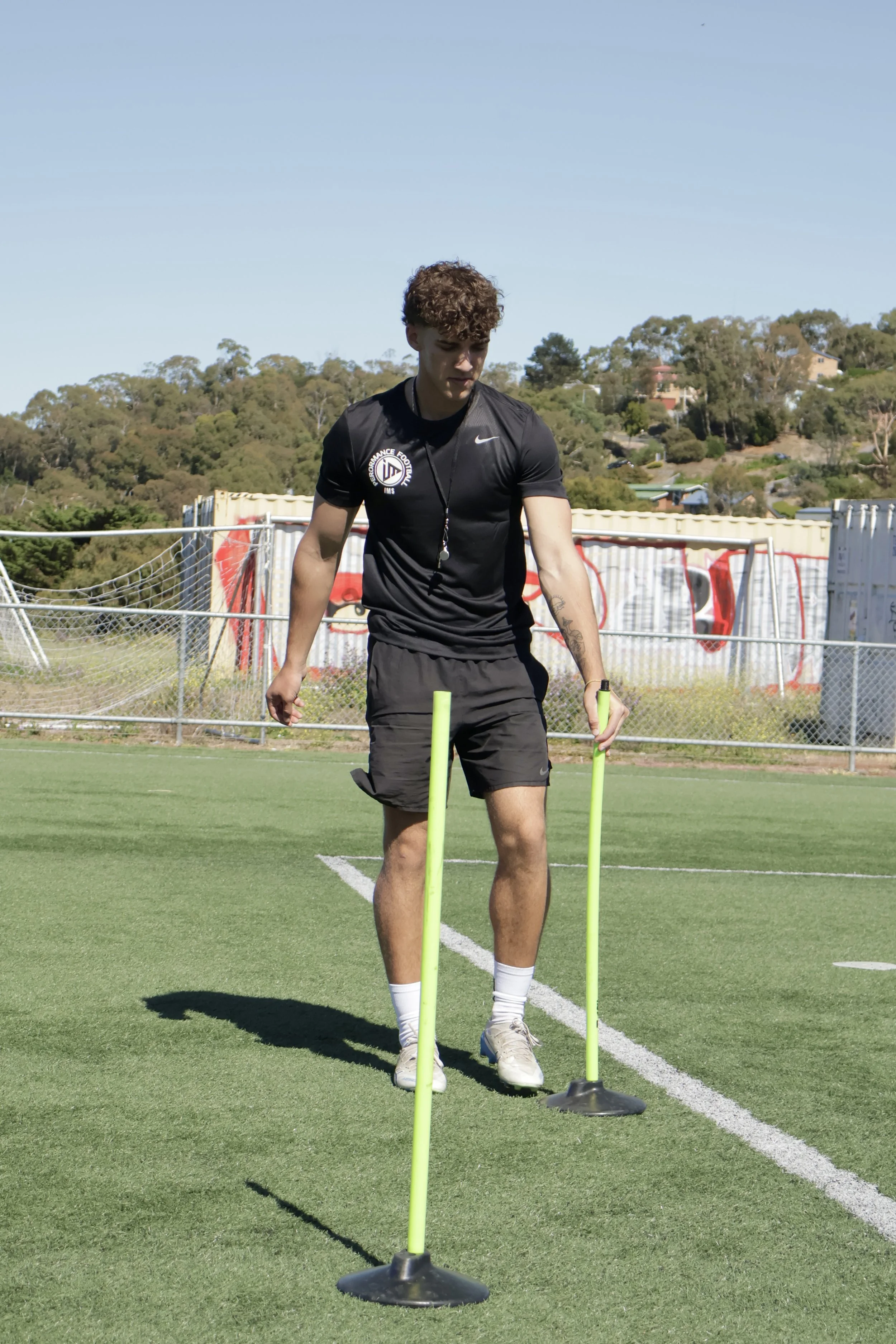 A young man in athletic clothing does agility drills with two green poles on a grassy field.