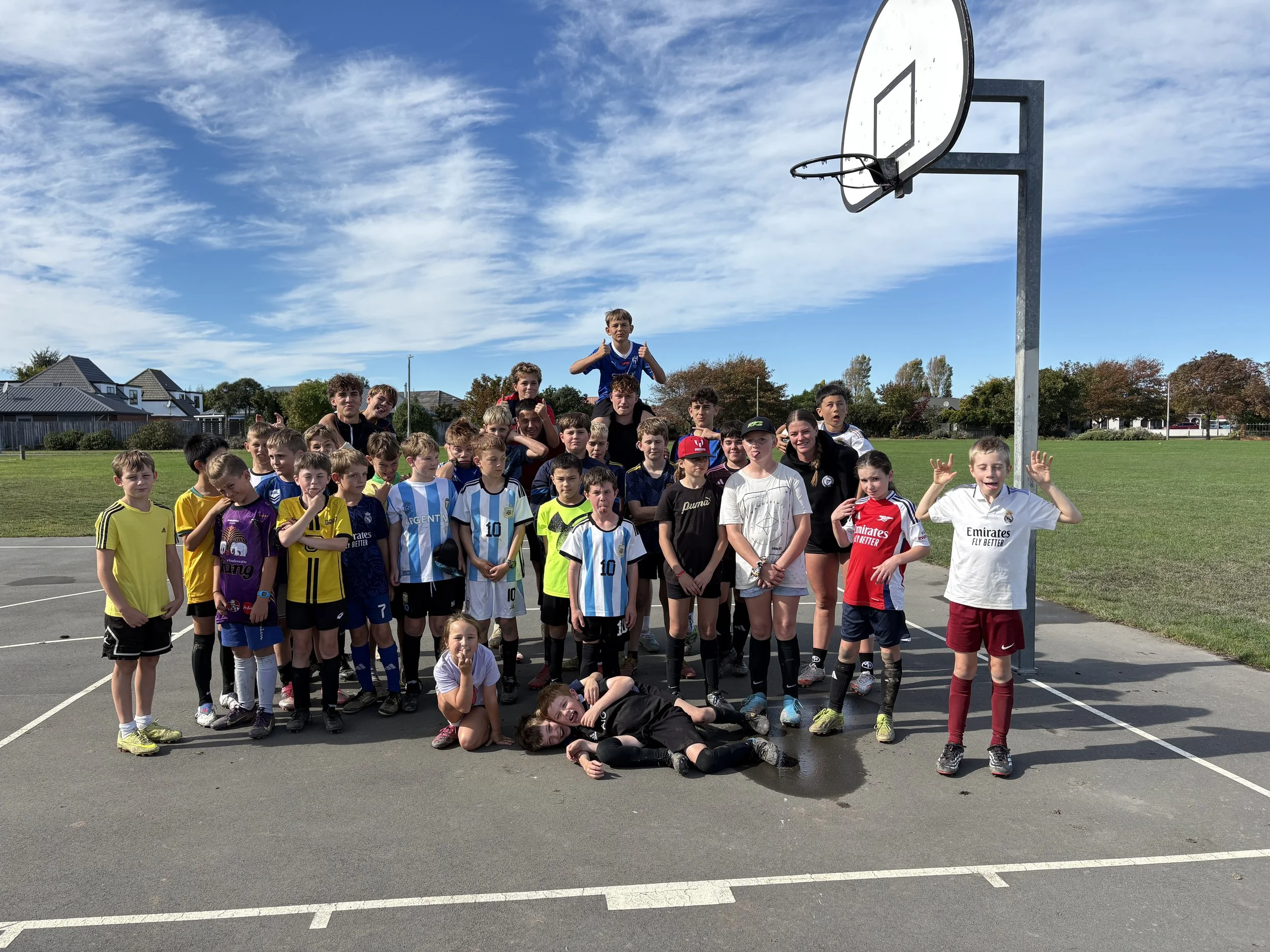 A large group of children and teenagers gathered outdoors on a basketball court, posing for a group photo. Some children are sitting or lying on the ground, while others stand behind. Two boys are sitting in front, making peace signs. In the background, there is a basketball hoop and a row of houses with trees. The sky is partly cloudy with patches of blue.